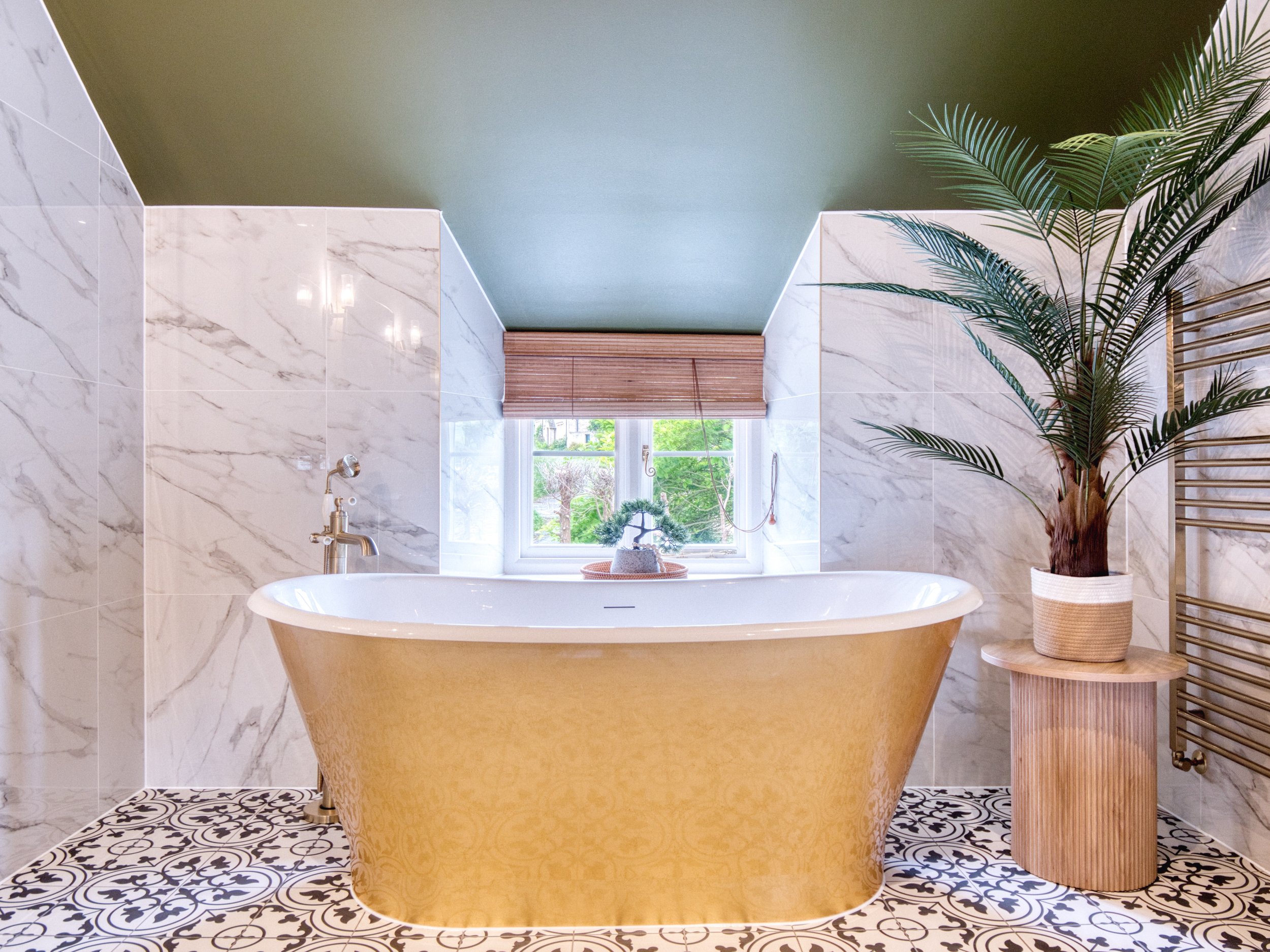 A bathroom with a freestanding bathtub with a gold exterior and patterned gold interior, a window with a bamboo shade, and a tall green indoor plant in a basket. The walls and ceiling are tiled with white marble tiles and a matte green paint, respectively. There is a small wooden side table next to the bathtub and patterned black and white floor tiles.