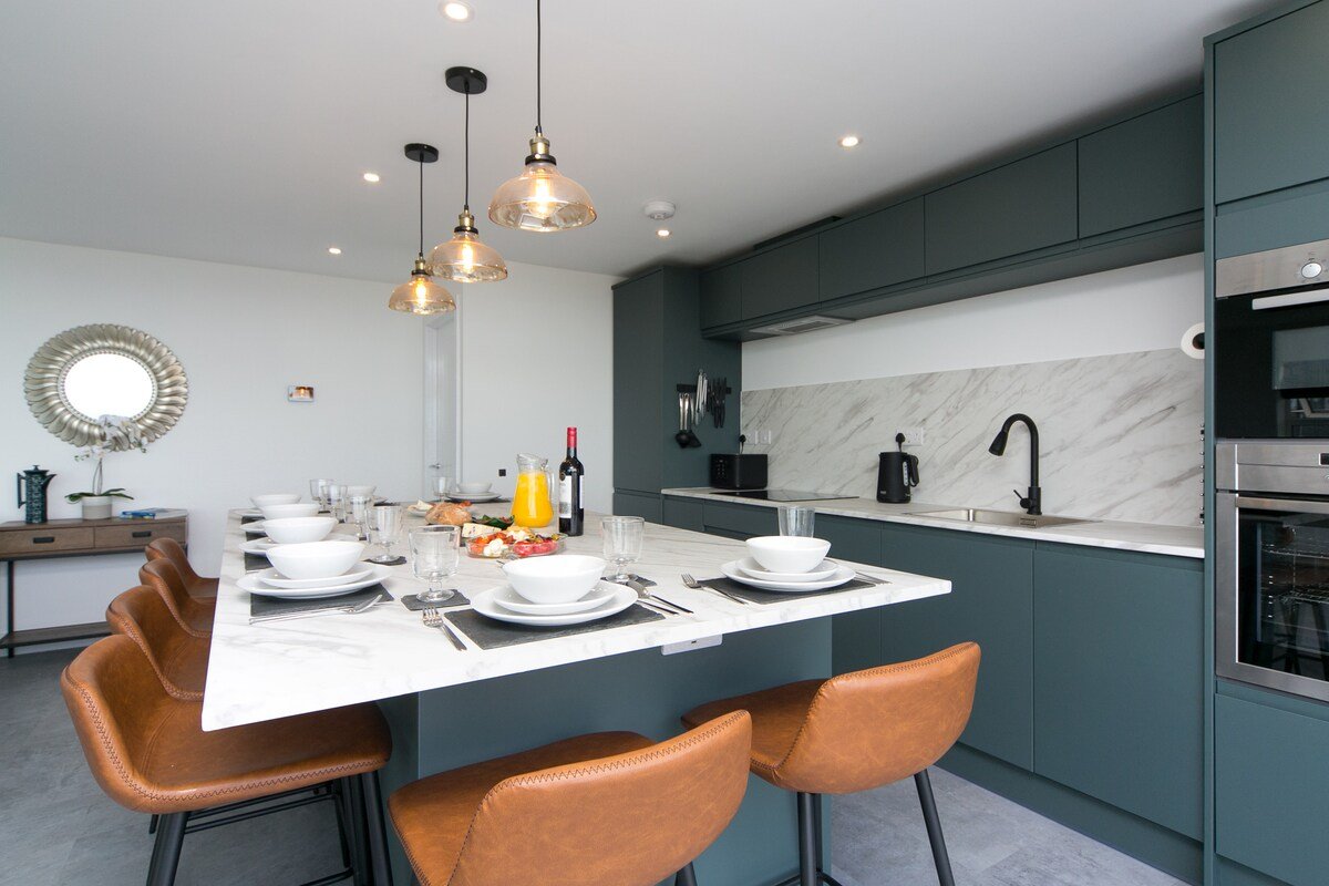 Modern kitchen with dark green cabinets, a white marble backsplash, and a white marble island set for a meal with plates, bowls, glasses, and silverware. There are three pendant lights above the island, and a few ceiling lights. Kitchen appliances include a built-in oven and microwave. Brown chairs are around the kitchen island.