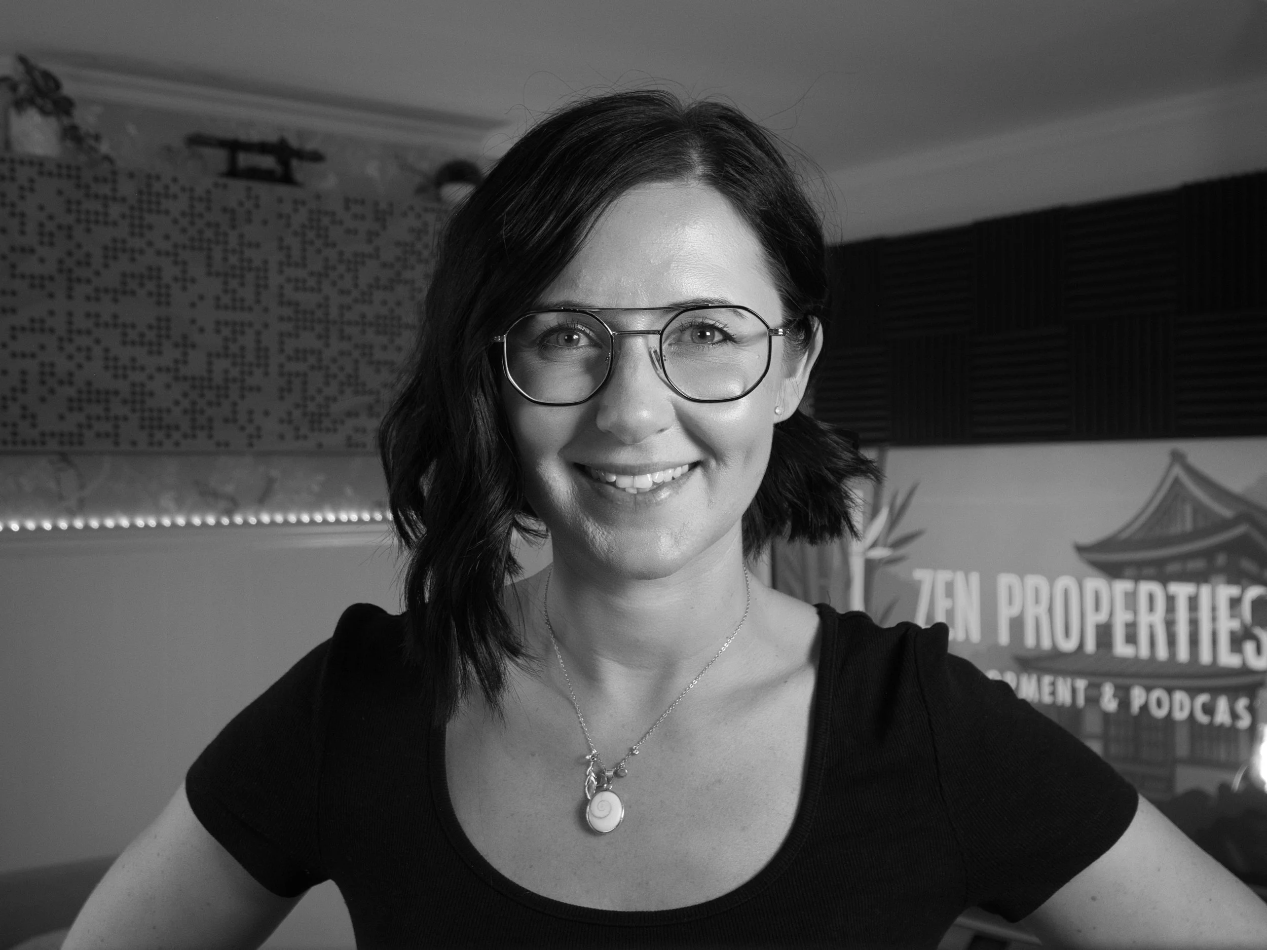 A smiling woman with glasses, dark hair, wearing a necklace with a snail pendant, and a black top, in front of a poster and an Asian-style building in the background.