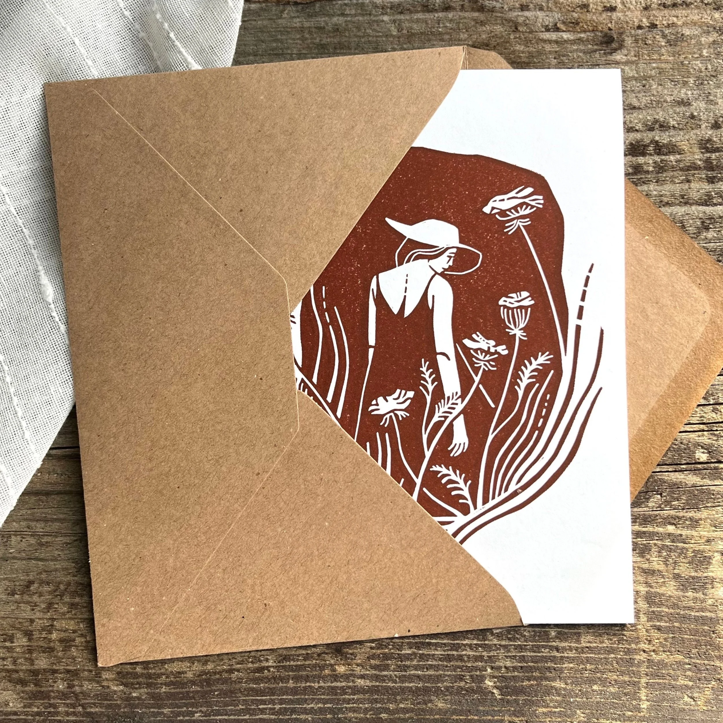 Karte mit einem Druck von einer Frau im Kleid mit Hut in einem Feld mit hohen Gräsern; in einem braunen Umschlag auf einem Holztisch.