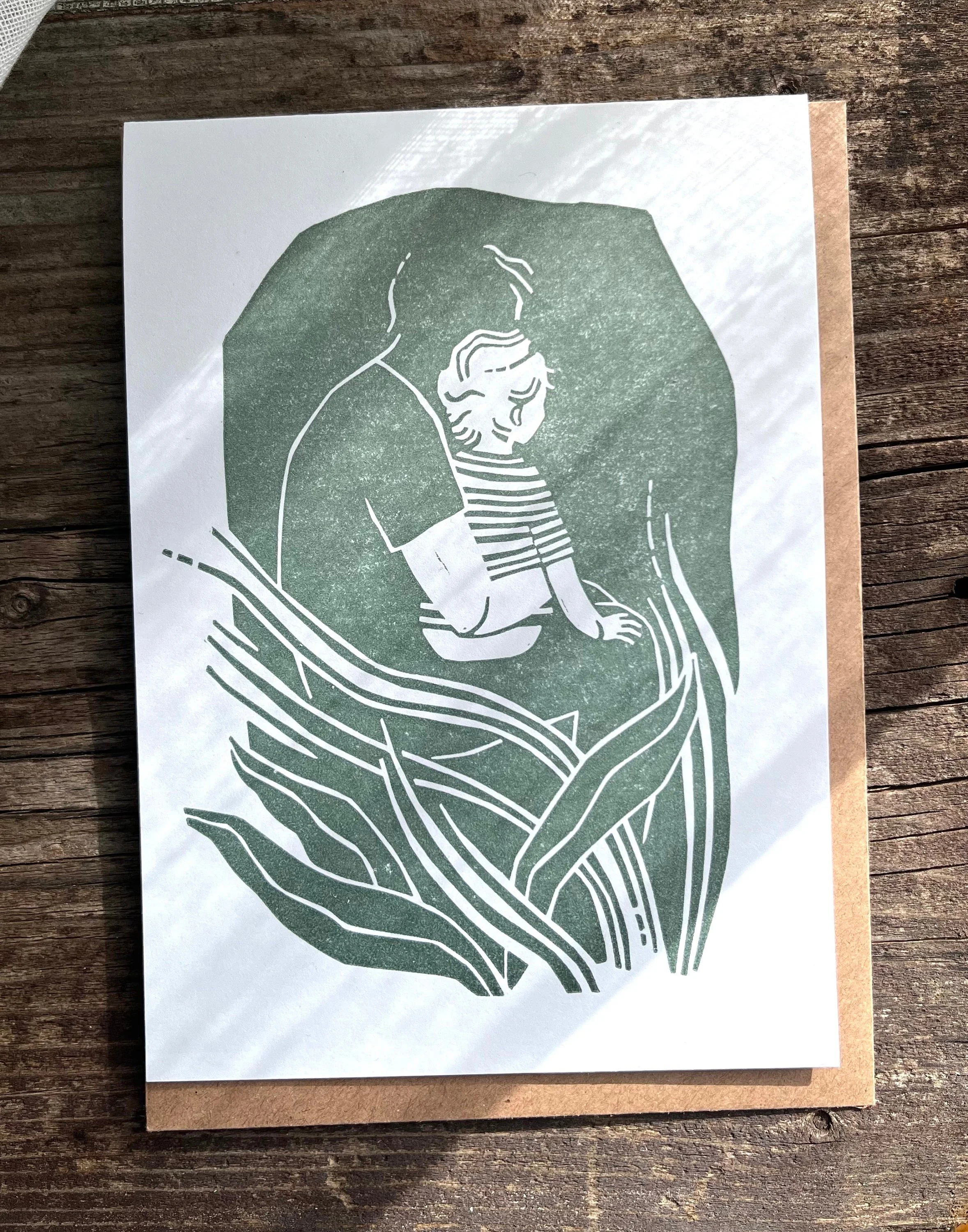 Linolschnitt eines Erwachsenen, der ein Kind hält, umgeben von stilisierten Pflanzen, auf einer Holzoberfläche liegend.