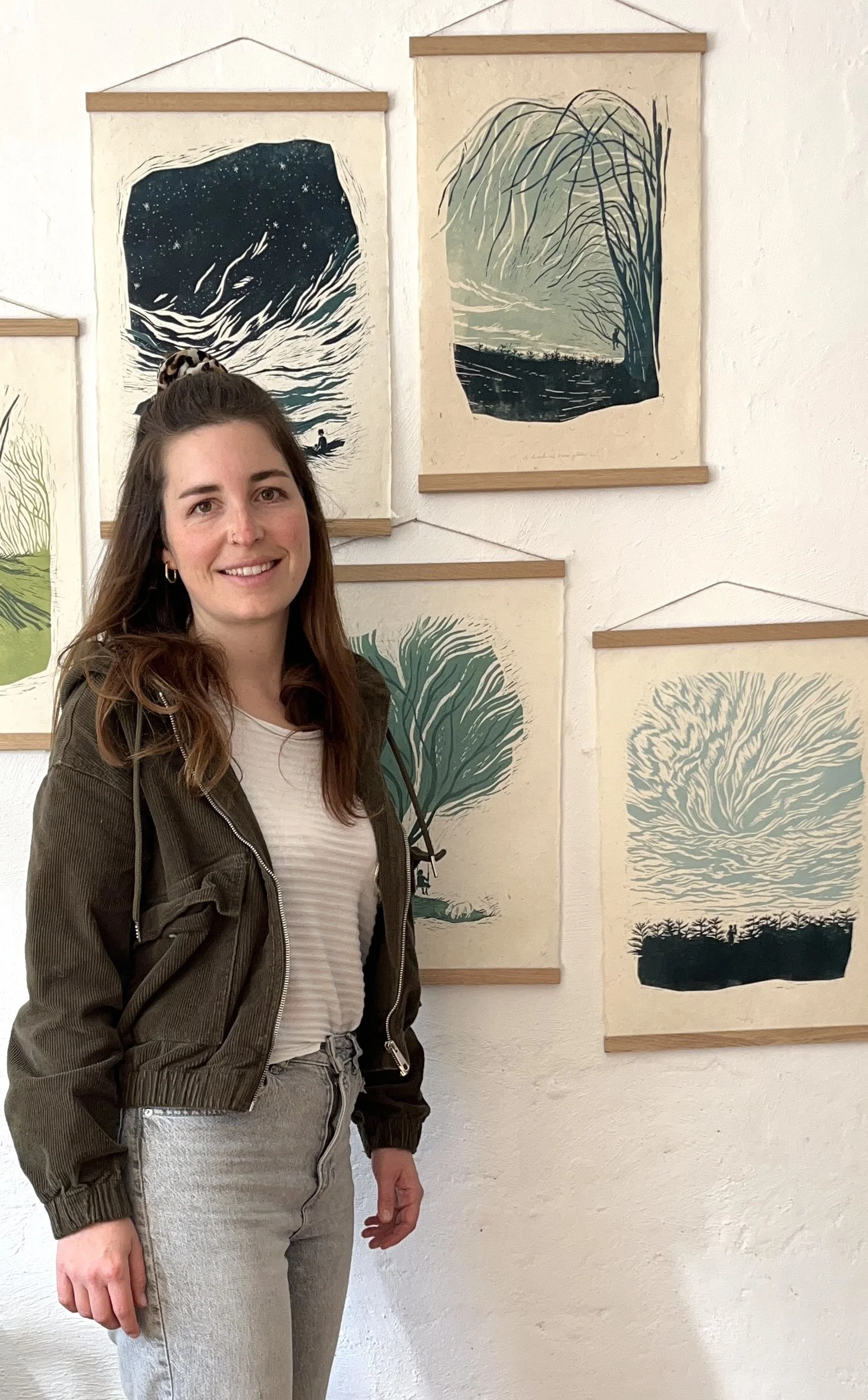 Eine Frau steht vor einer Wand mit mehreren gerahmten, künstlerischen Drucken von Bäumen und Naturmotiven. Sie trägt eine grüne Jacke über einem weißen gestreiften T-Shirt und Jeans.