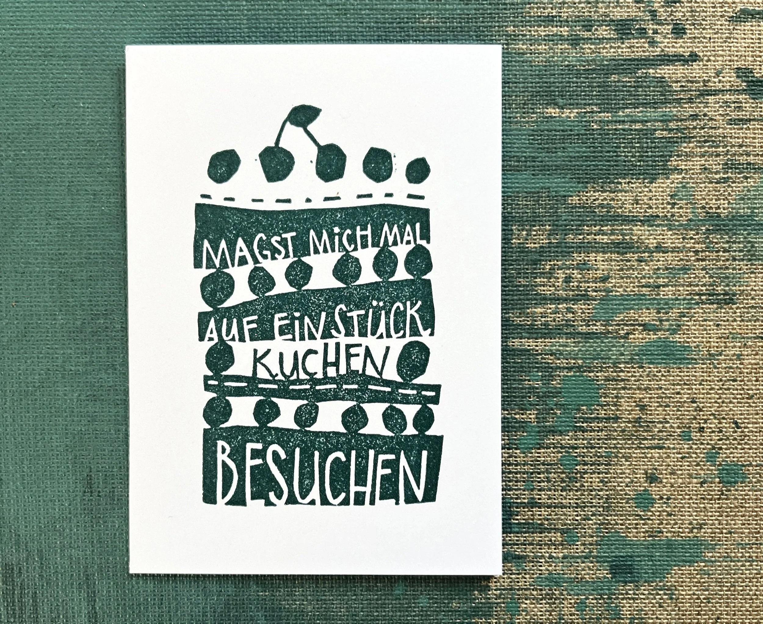 Handgemachte Grußkarte mit dem Text: 'Magst mich mal auf ein Stück Kuchen besuchen', dekoriert mit stilisierten Kirschen darüber. Hintergrund aus grünem Stoff mit unregelmäßigem Muster.