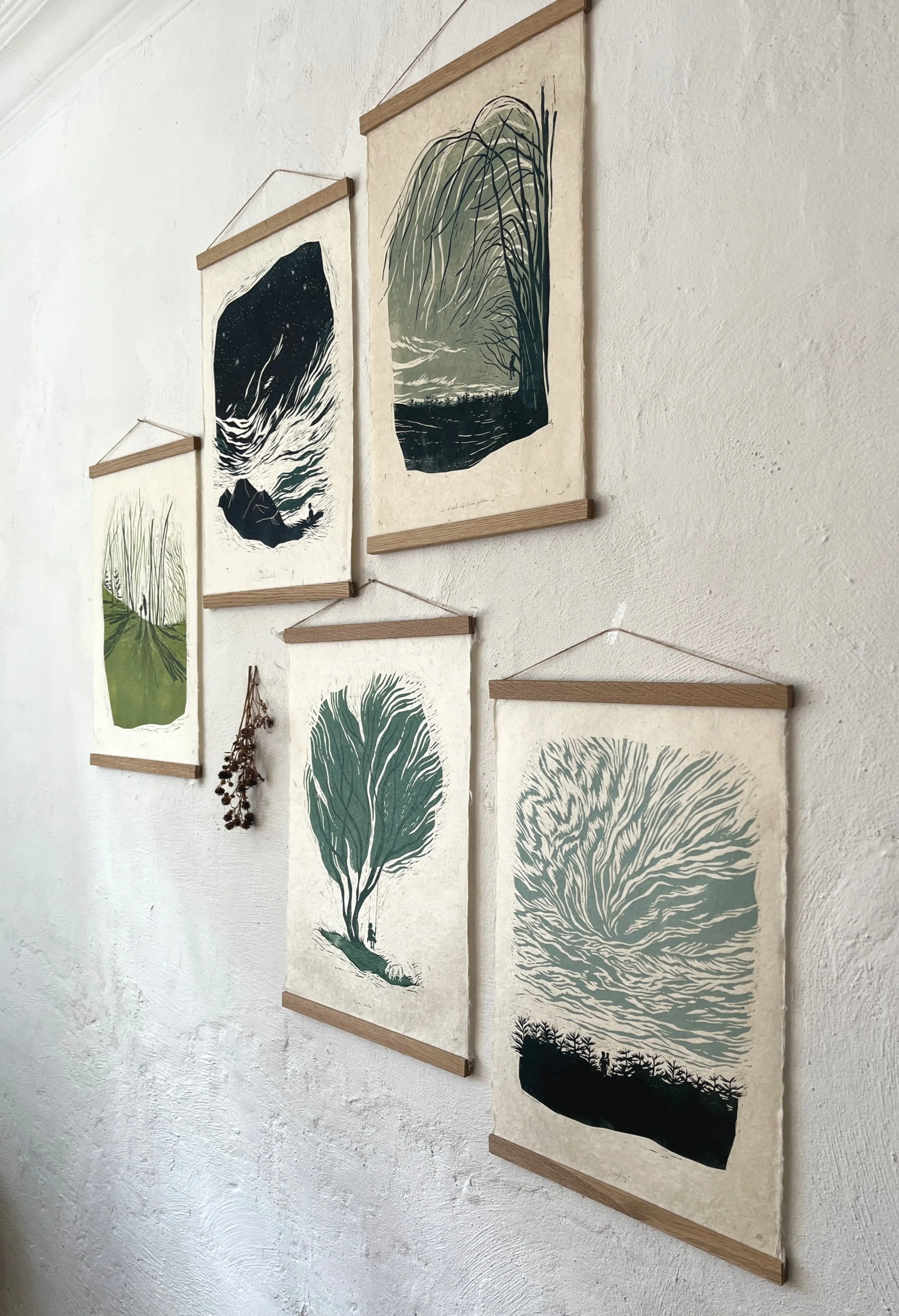 Fünf Wandposter mit Landschaftsillustrationen in Grün, Schwarz und Grau, an einer weißen Wand, dekoriert mit getrockneten Blumen.