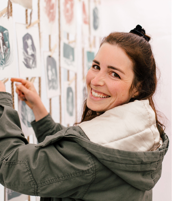 Eine lächelnde Frau in einer grünen Jacke betrachtet und arrangiert Kunstkarten an einer Wand.