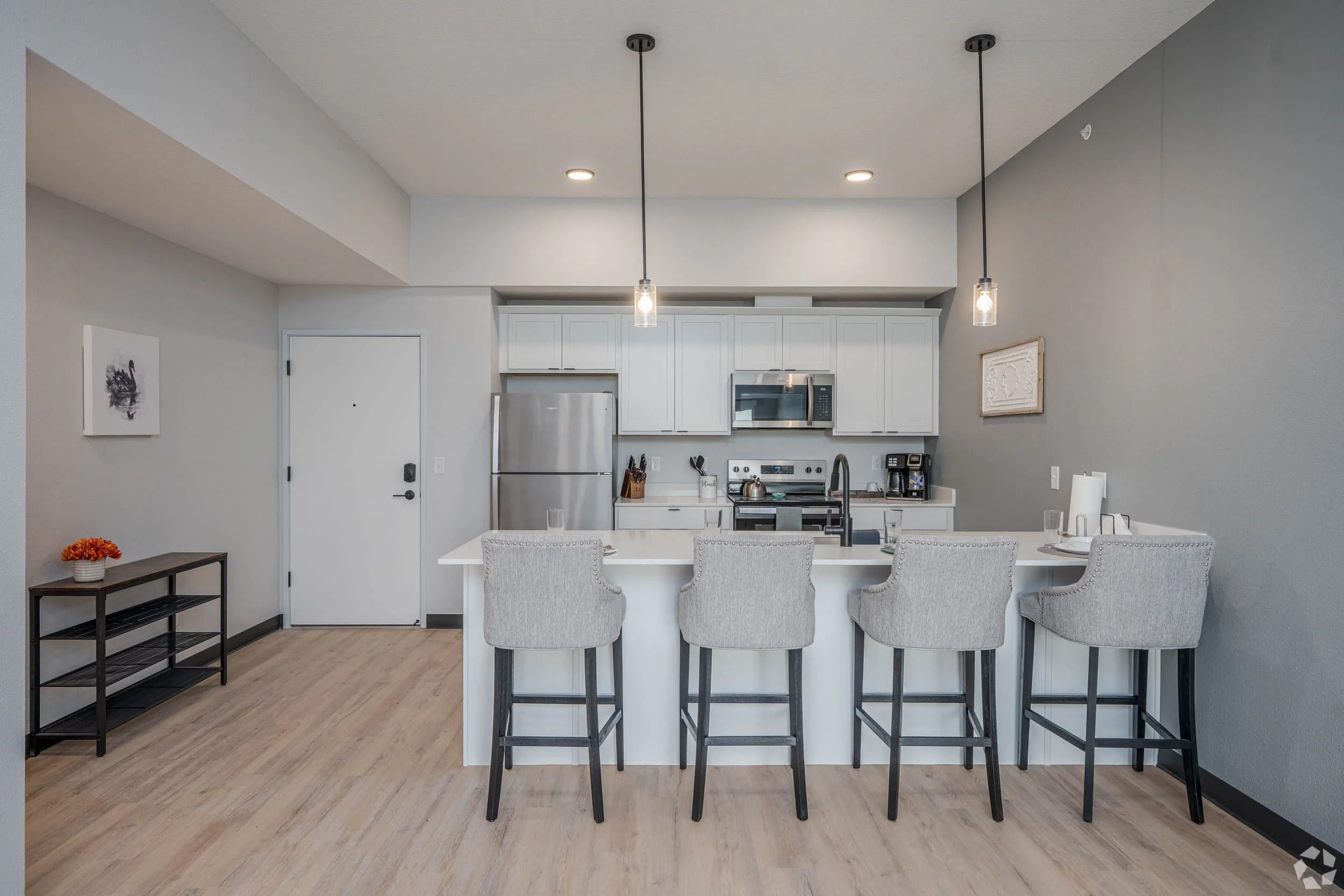 Modern kitchen in apartments in Rochester with white cabinets, stainless appliances, and a white island with barstools under pendant lights.