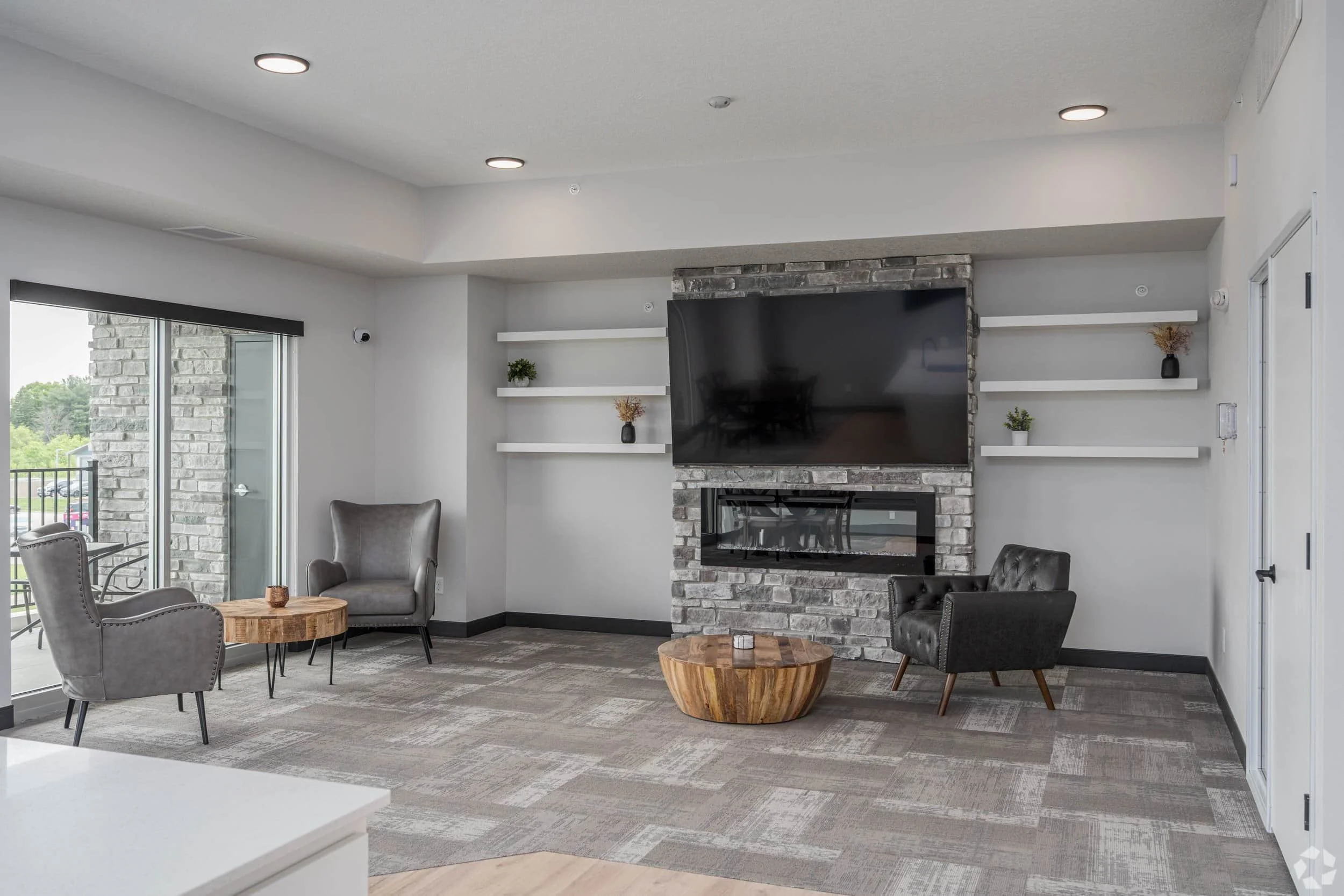 Modern lounge in apartments in Rochester with gray chairs, TV above brick fireplace, shelves with decor, and round wood tables.