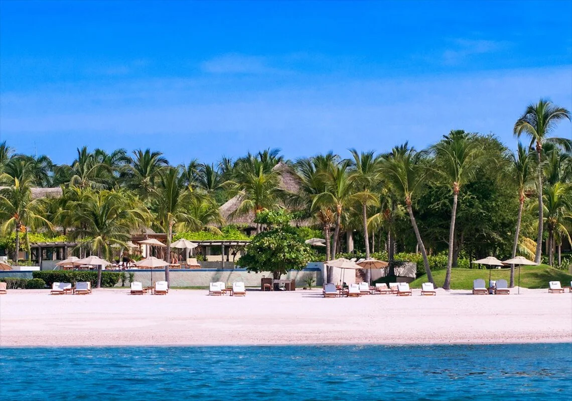 A tropical beach with white sand, lounge chairs, umbrellas, and lush palm trees under a bright blue sky, with calm ocean water in the foreground.