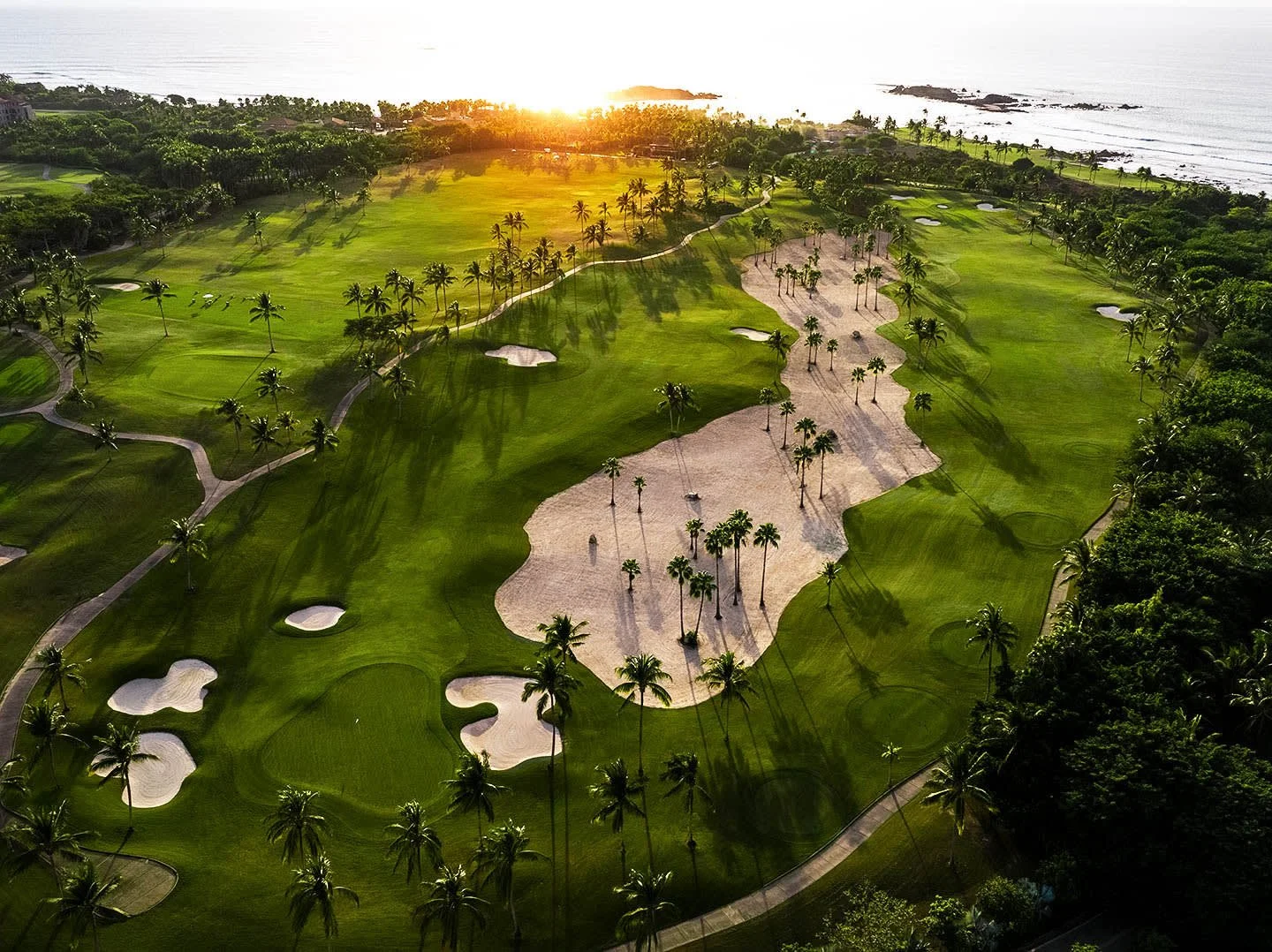 Aerial view of a tropical golf course with palm trees, sand traps, and lush green fairways near the ocean at sunset.