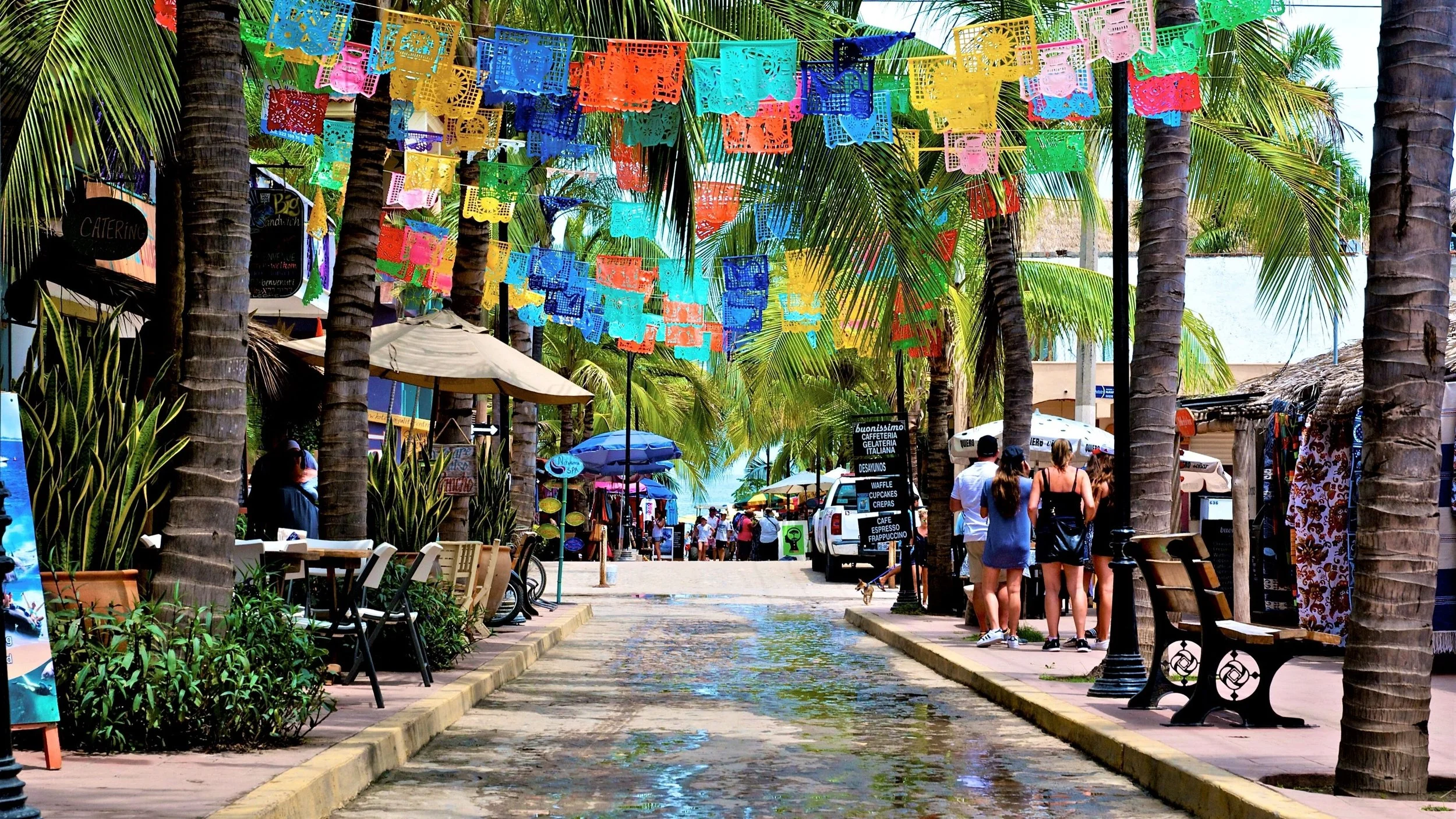 Colorful street with papel picado banners, palm trees, shops, and people walking.