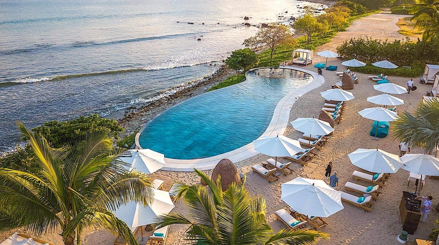 Aerial view of a beachside pool area with lounge chairs and umbrellas, overlooking the ocean and sandy shore.