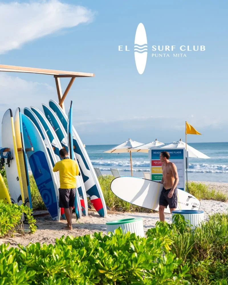 Beach scene at El Surf Club, Punta Mita, with surfboards and people preparing for surfing near the ocean under sunny skies.