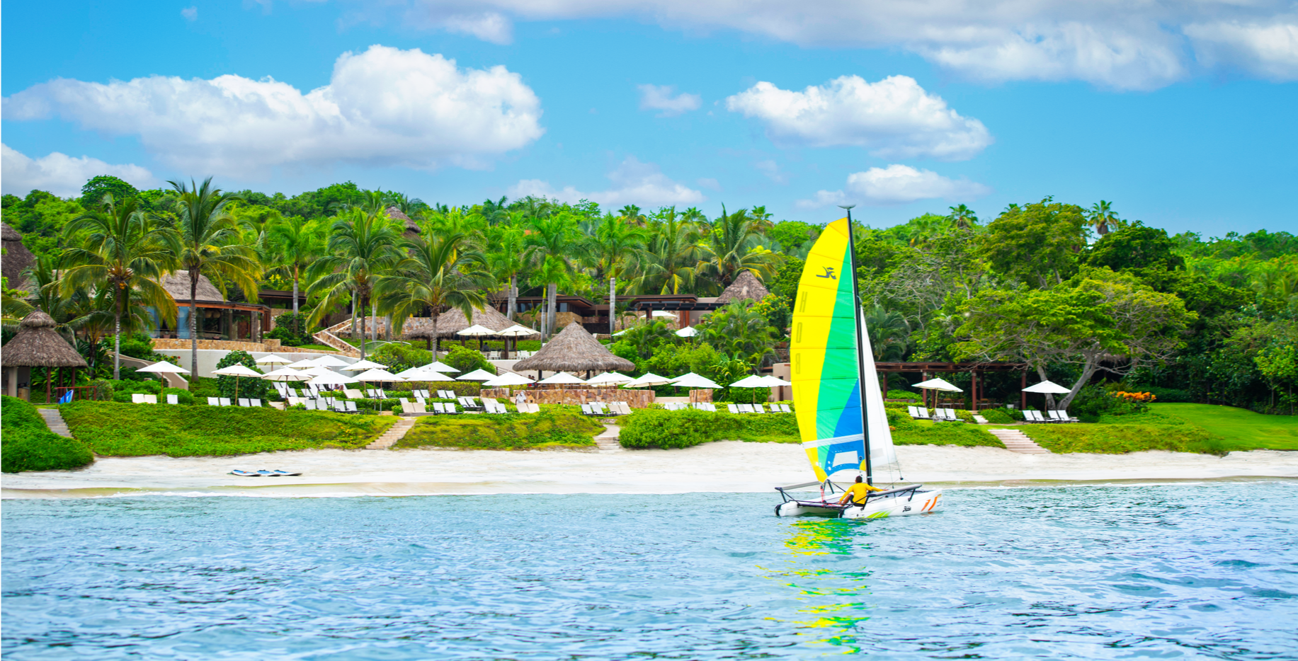 Beach resort with a yellow sailboat on the water, lush greenery, palm trees, and white umbrellas.