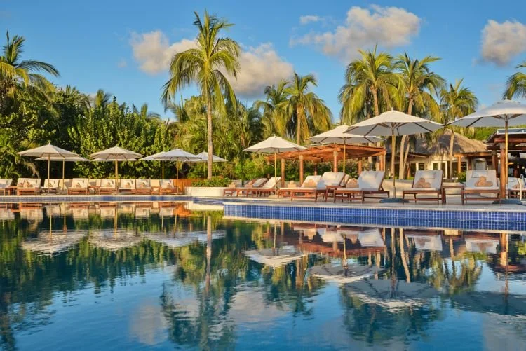 Tropical poolside scene with lounge chairs, umbrellas, palm trees, and a clear blue sky