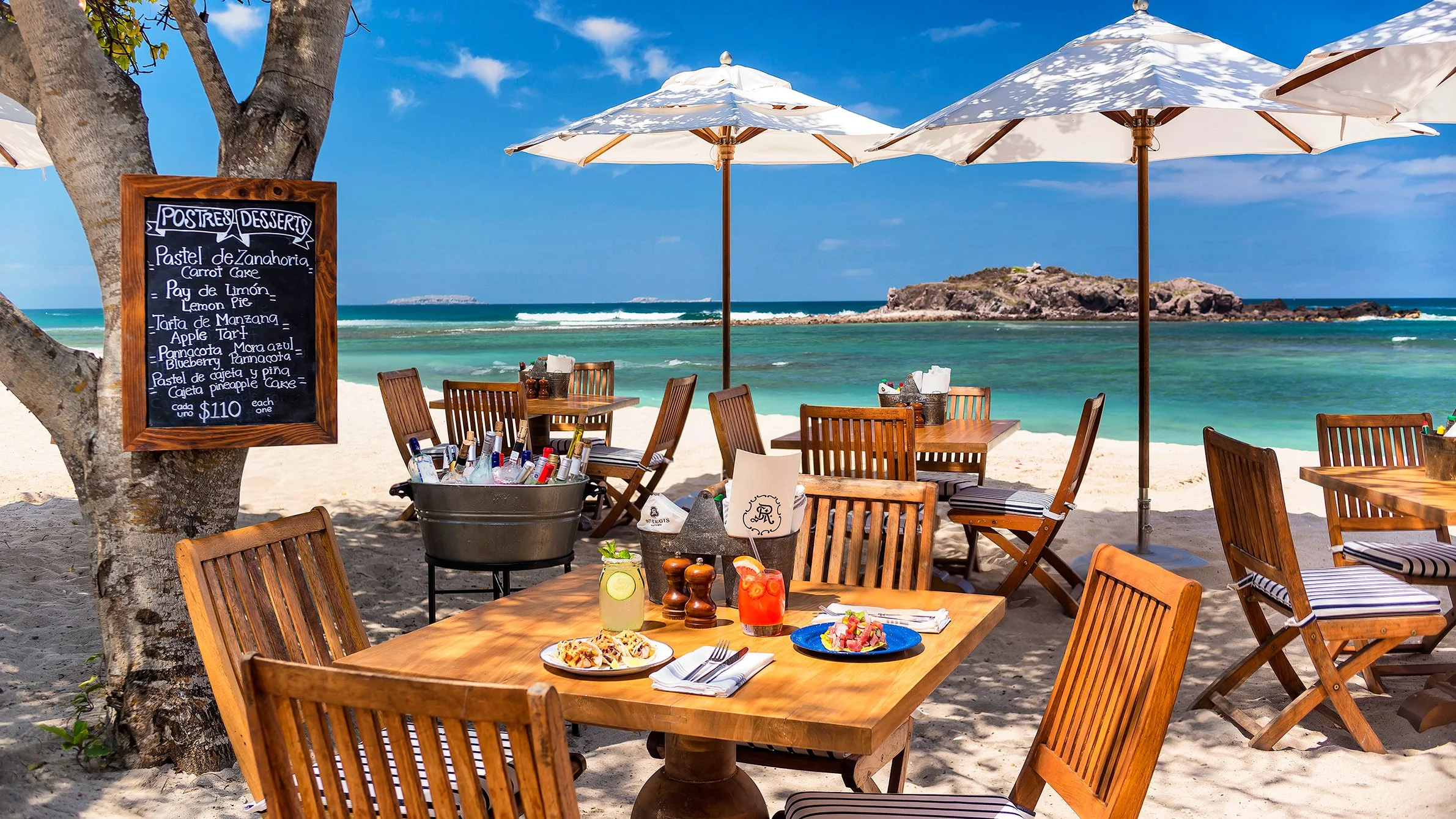 Beachside restaurant with wooden tables and chairs under white umbrellas, overlooking the ocean with a small island or rock formation in the distance.