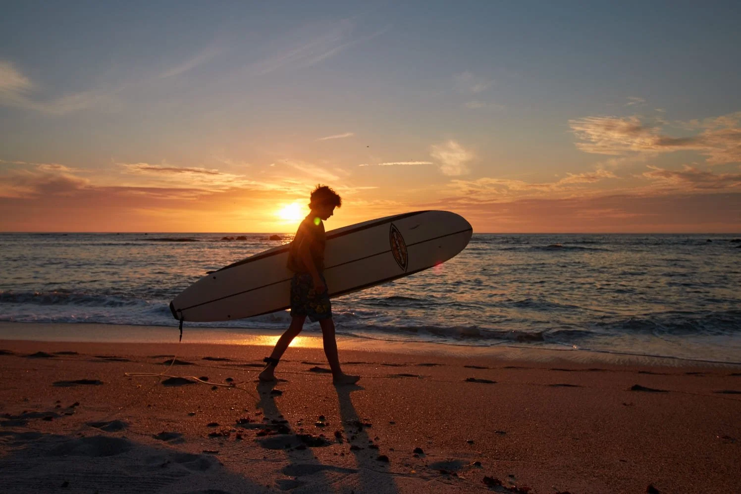 Silhouette of a person walking on the beach carrying a surfboard at sunset.