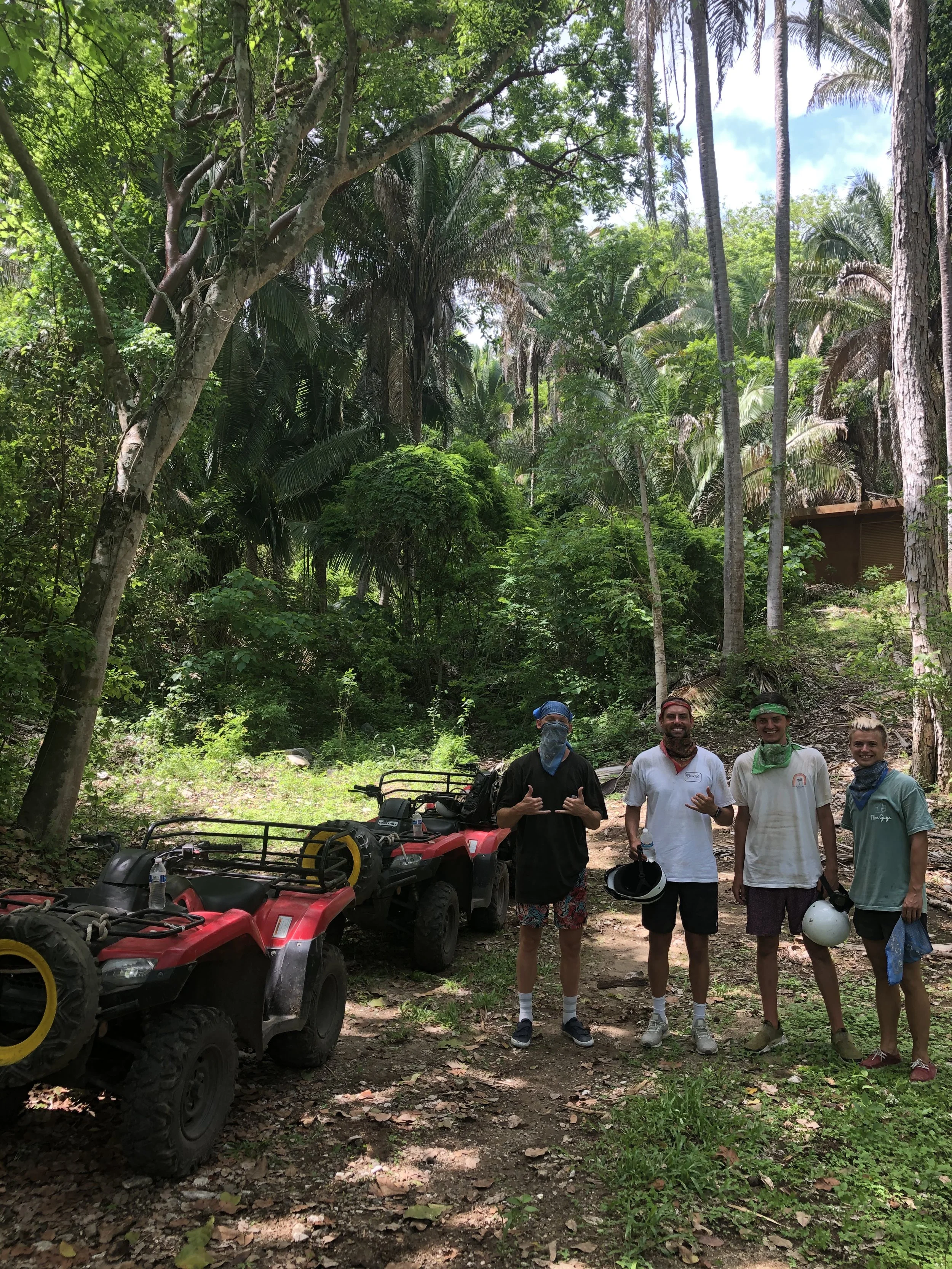 Four people standing in a forested area next to two red ATVs, wearing casual clothing and bandanas, holding helmets, surrounded by trees and greenery.