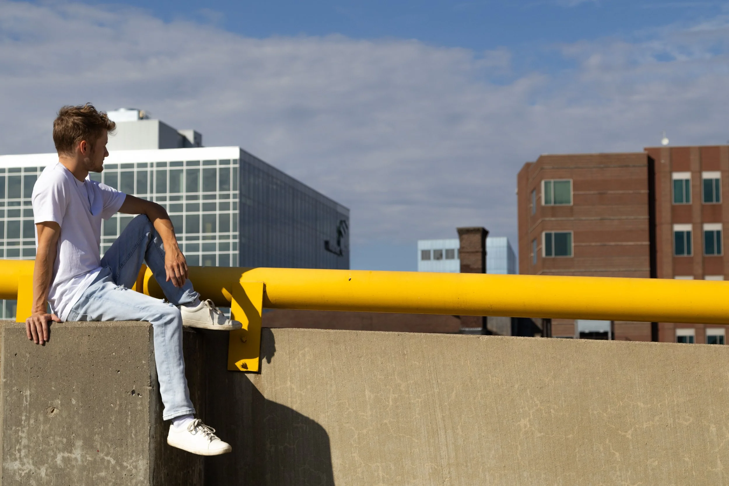 A young man in a white t-shirt and light blue jeans sitting on a concrete ledge next to a yellow railing in an urban area, with modern buildings and a partly cloudy sky in the background.