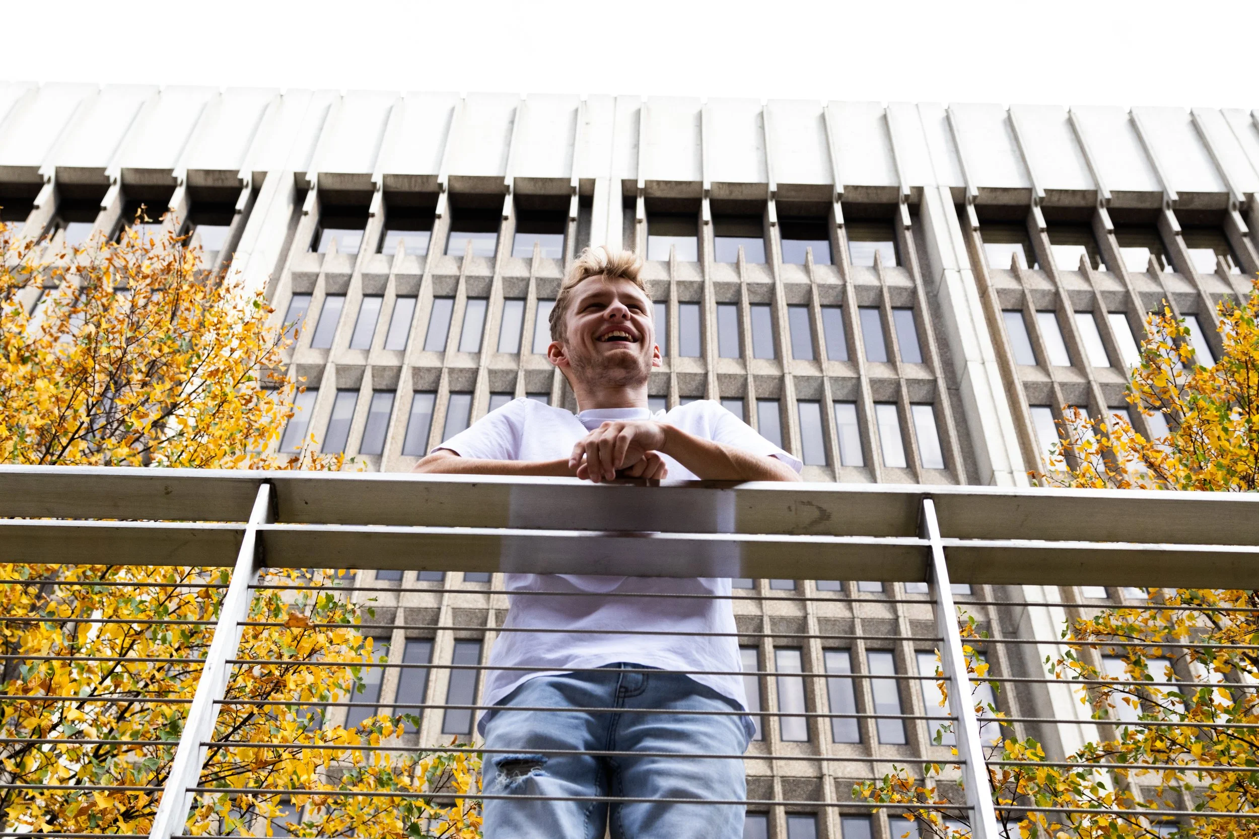 Young man leaning on a metal railing, smiling, in front of a large concrete building with rectangular windows, surrounded by autumn-colored trees.