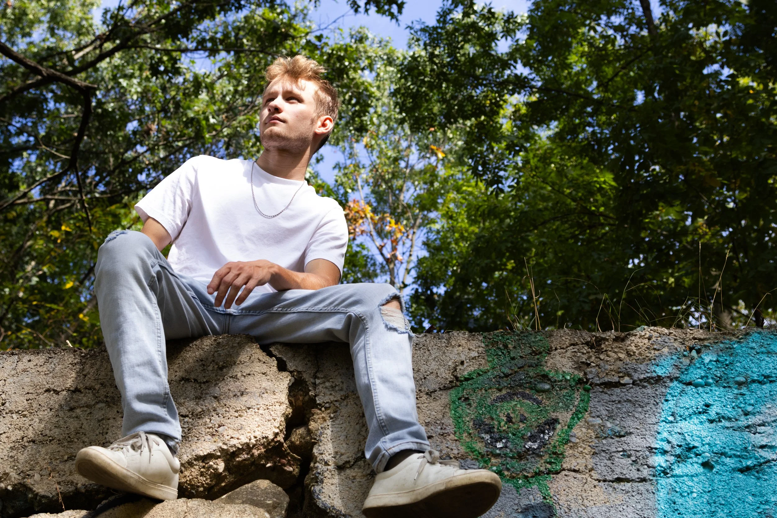 Young man sitting on a concrete wall with graffiti, wearing a white T-shirt, ripped jeans, and white sneakers, outdoors with green trees and blue sky in the background.
