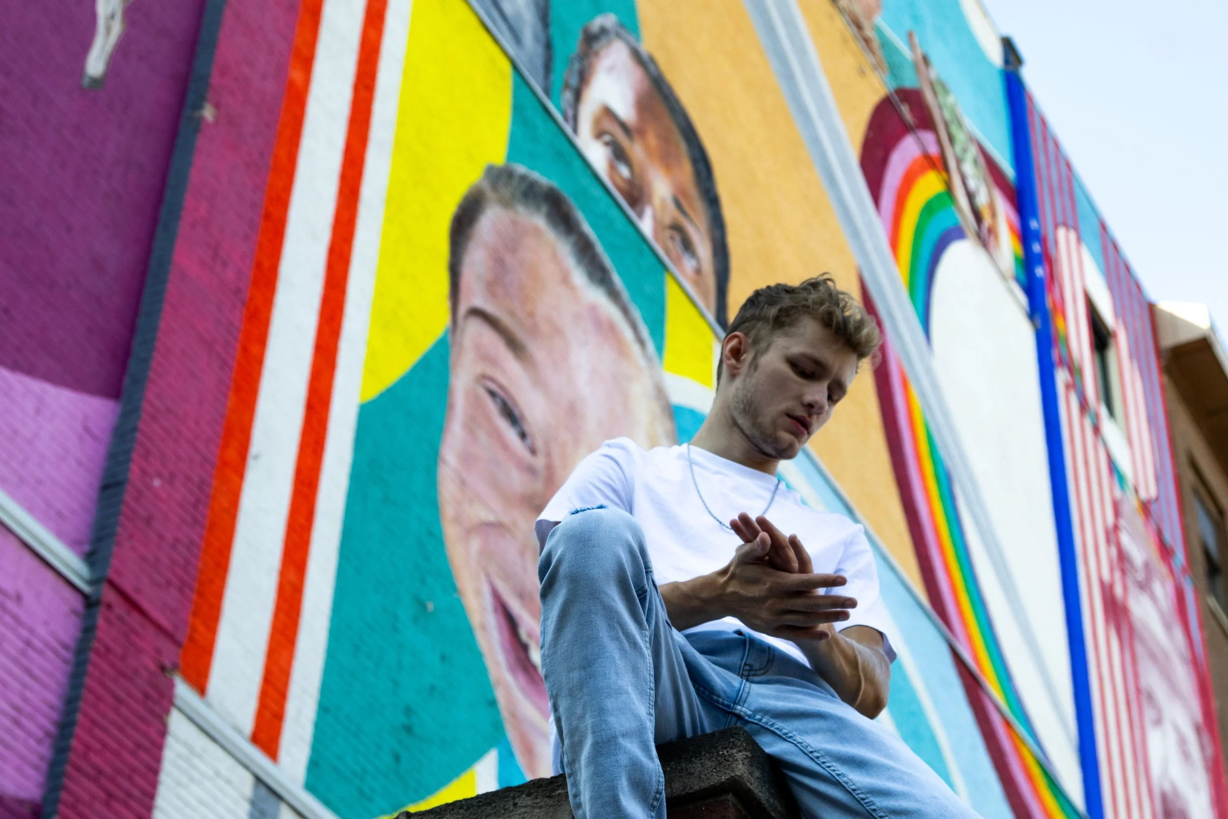 A young man sitting on a ledge in front of a colorful mural featuring large portraits of diverse faces and rainbow designs, looking down at his hands.