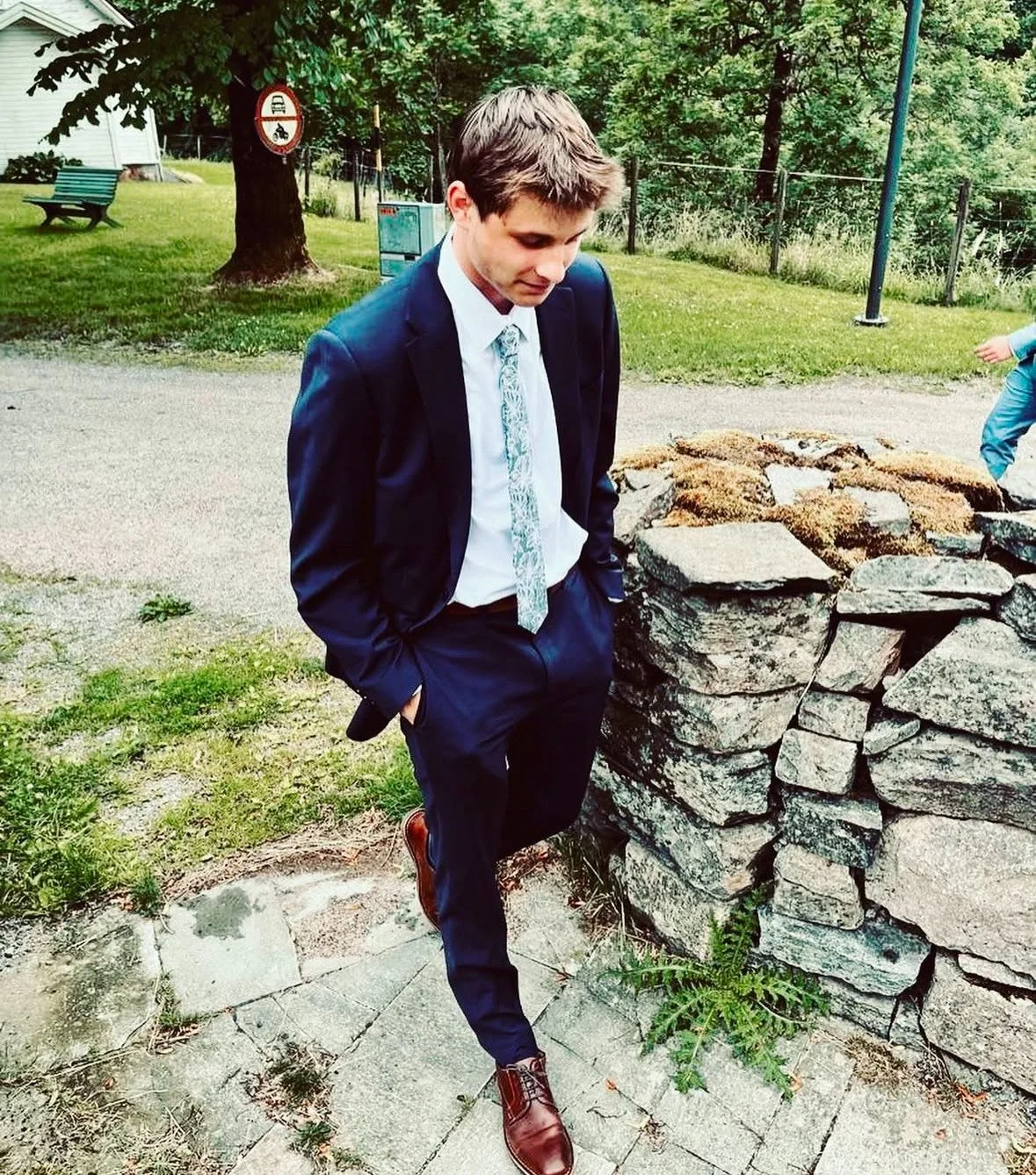 A young man in a blue suit with a patterned tie stands with hands in pockets next to a rocky stone structure outdoors in a park, with trees, a bench, and a person in the background.