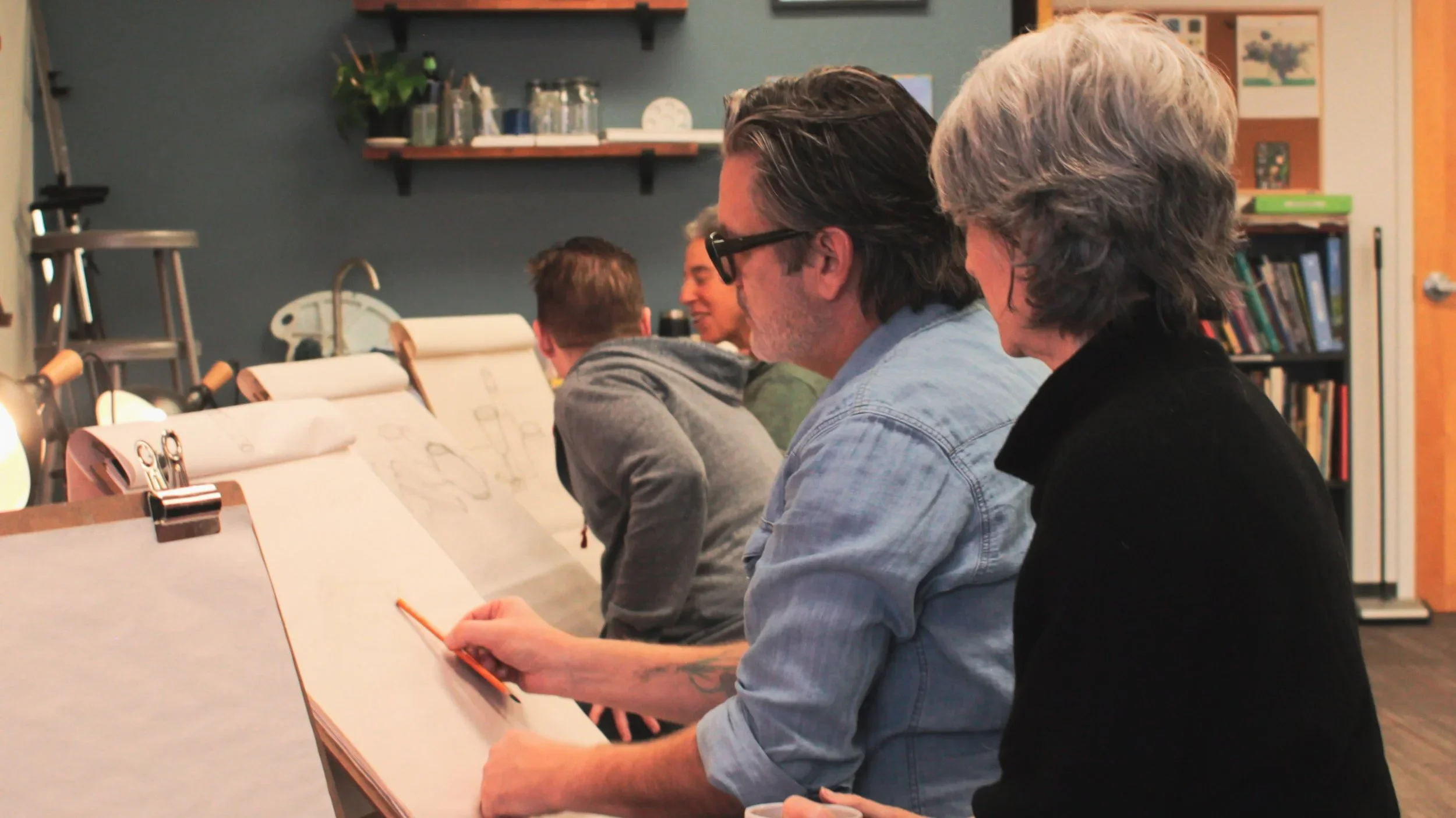 A group of people seated at a table in a classroom or workshop setting, engaging with sketches or drawings on clipboards, with art supplies and books in the background.
