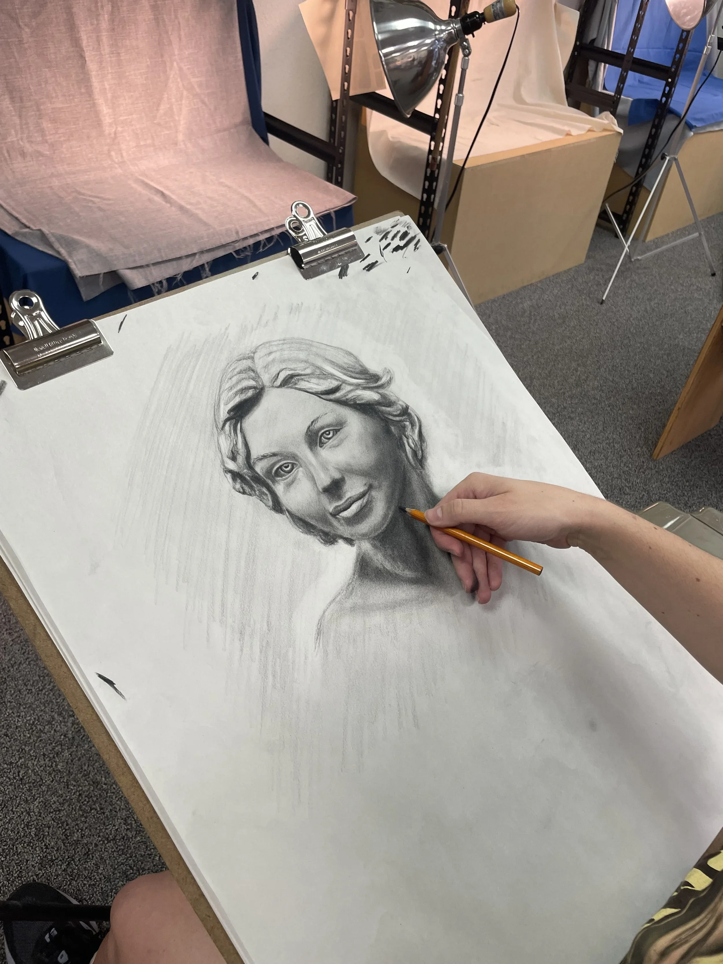 A person drawing a realistic pencil portrait of a woman with long wavy hair on a large white sheet of paper. The workspace includes a pink cloth draped over a chair and a lamp illuminating the drawing. Shelves and other art supplies are visible in the background.