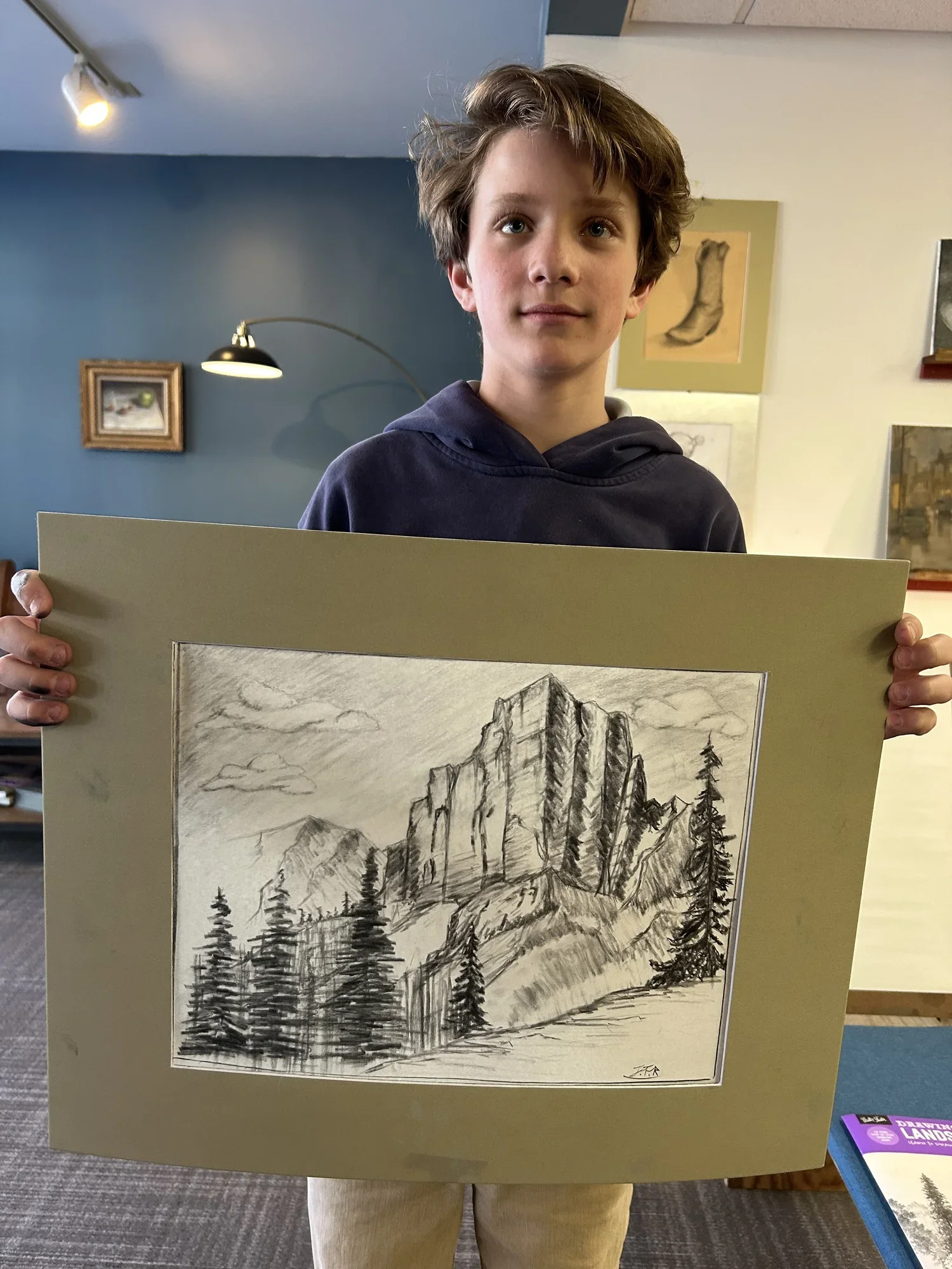 A boy with brown hair holding a piece of art depicting mountains, trees, and a cloudy sky in pencil sketch style.