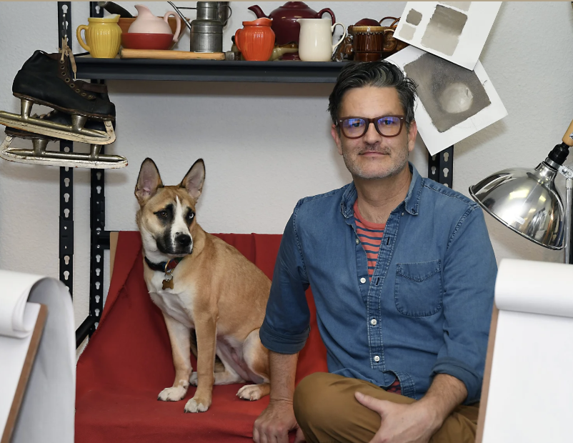 A man with dark hair and glasses sitting next to a tan and white dog on a red couch in an art studio surrounded by art supplies and equipment.