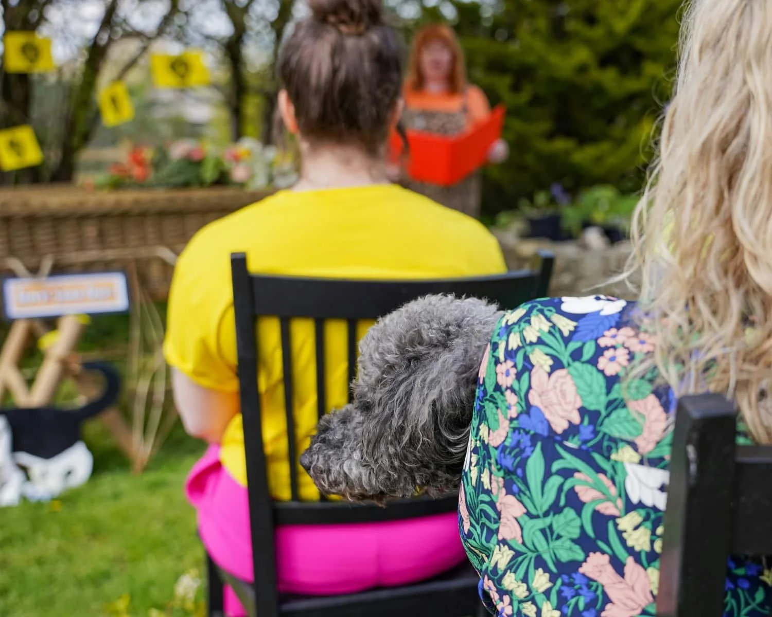 People seated outdoors at an informal memorial gathering, listening to a reading, with a dog resting its head on a chair.