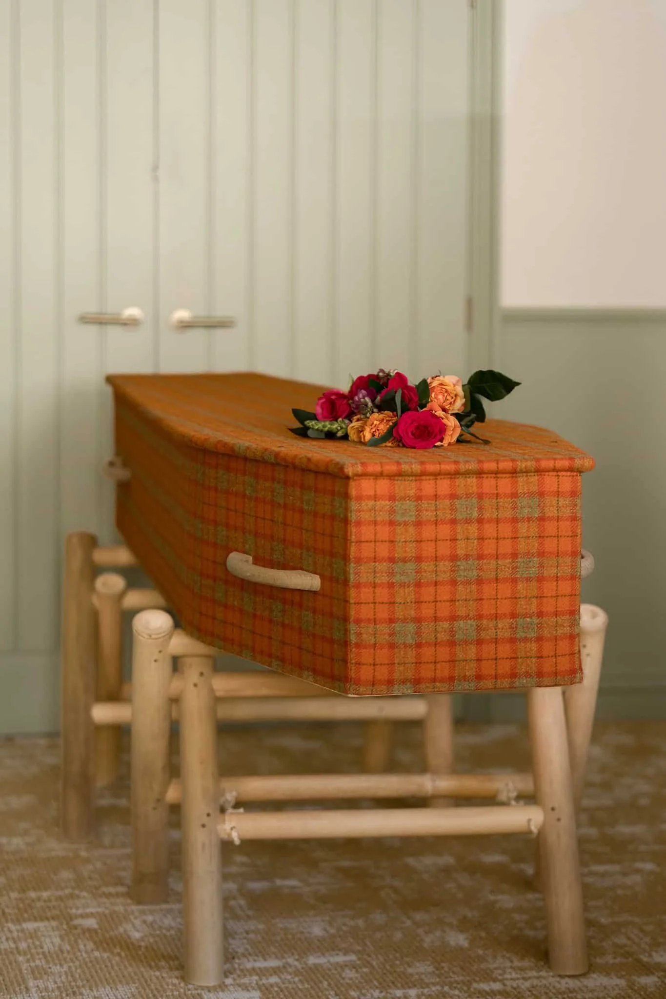 A wooden table with a red and orange plaid tablecloth, topped with a small bouquet of red, pink, and orange flowers with green leaves, in a room with pale green paneled walls and a whiteboard.