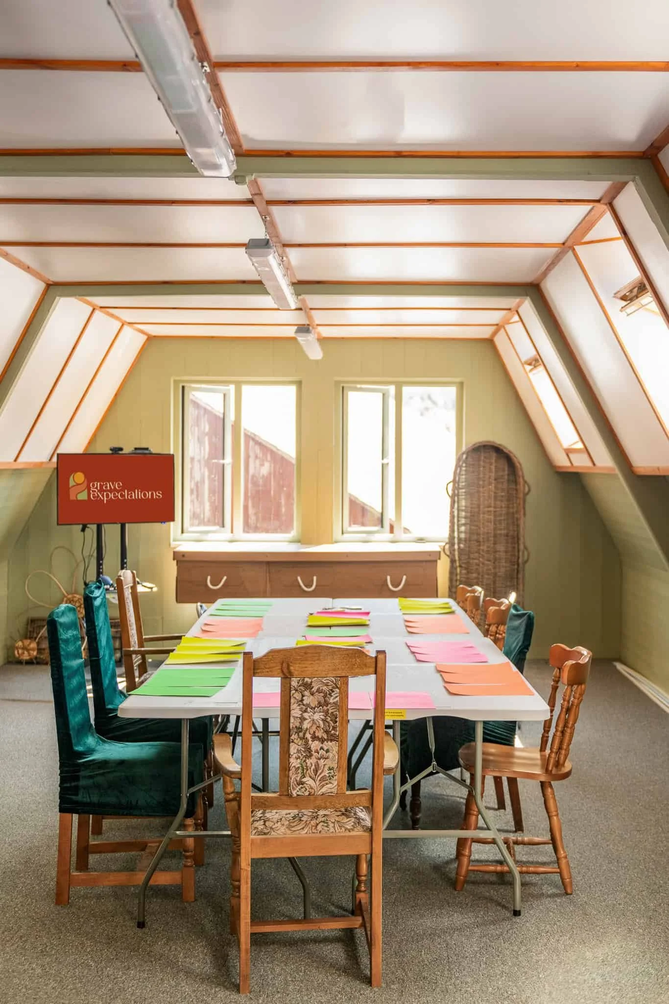 A room with a high, sloped ceiling and three large windows letting in natural light. There is a long table set with colorful paper and a mixture of wooden and upholstered chairs around it. In the background, a flat-screen TV displays the logo 'grave expectations' on a stand.