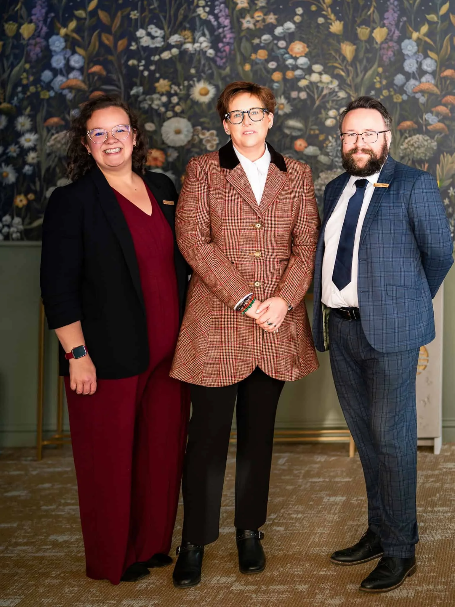 Three professionally dressed individuals standing side by side indoors, with a floral patterned wall behind them.