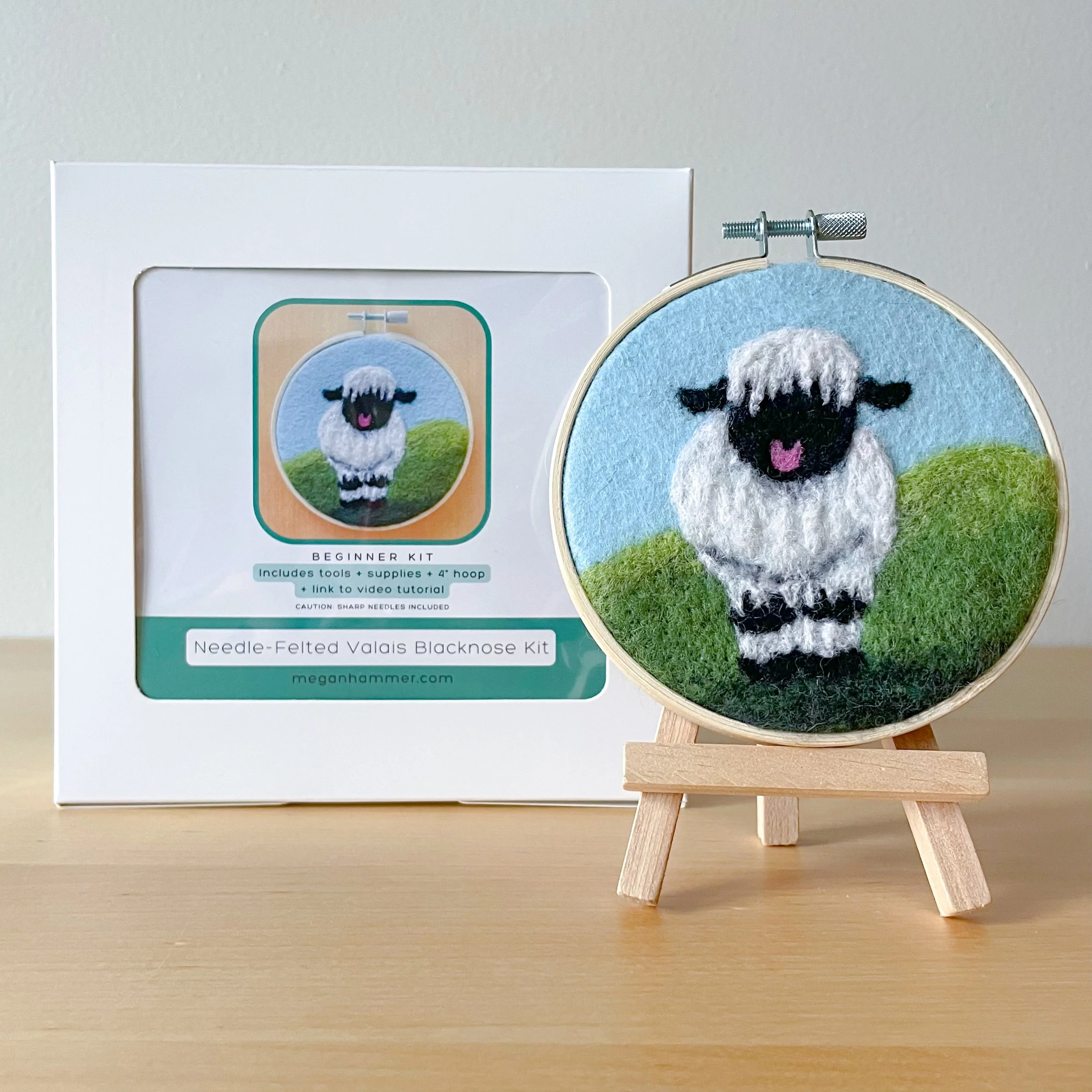 Felted Valais Blacknose Sheep art framed in an embroidery hoop sitting on a mini easel, in front of the white box of the kit.