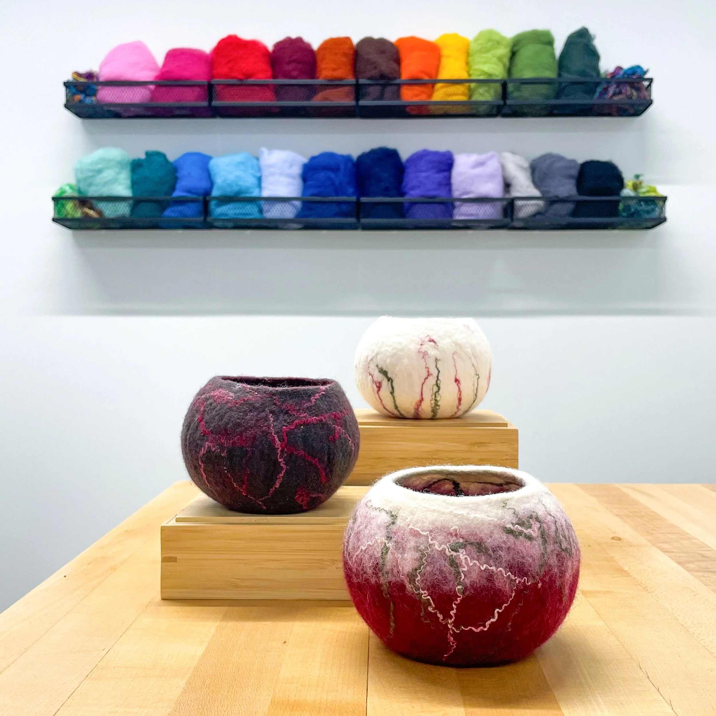 Three wet-felted bowls in white, dark green, and maroon with white and green accents sit on a light wood table with a display of colorful wool on the wall behind.