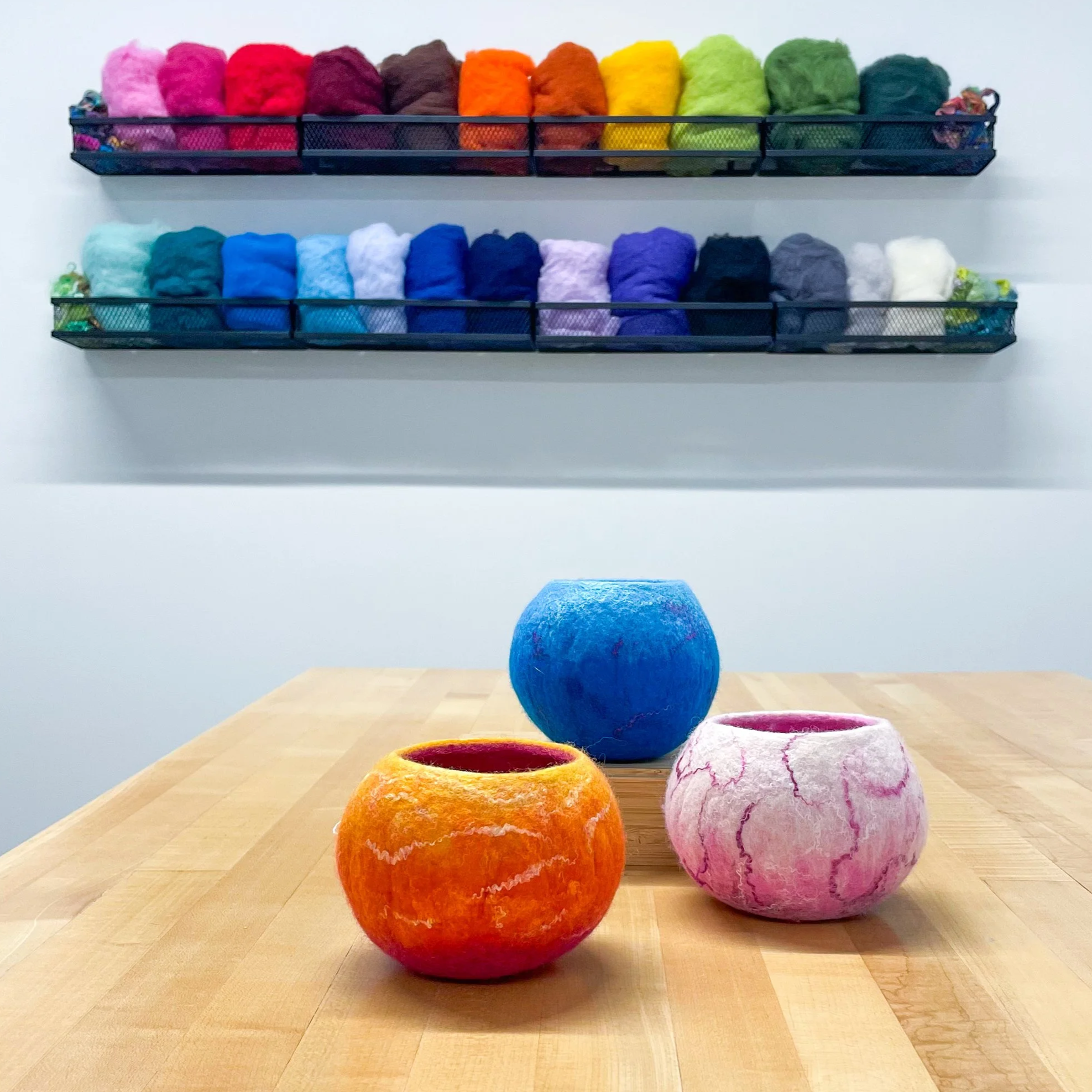 Three colorful felt bowls in shades of orange, blue, and pink sit on a light wood table with a display of colorful wool on the wall behind.