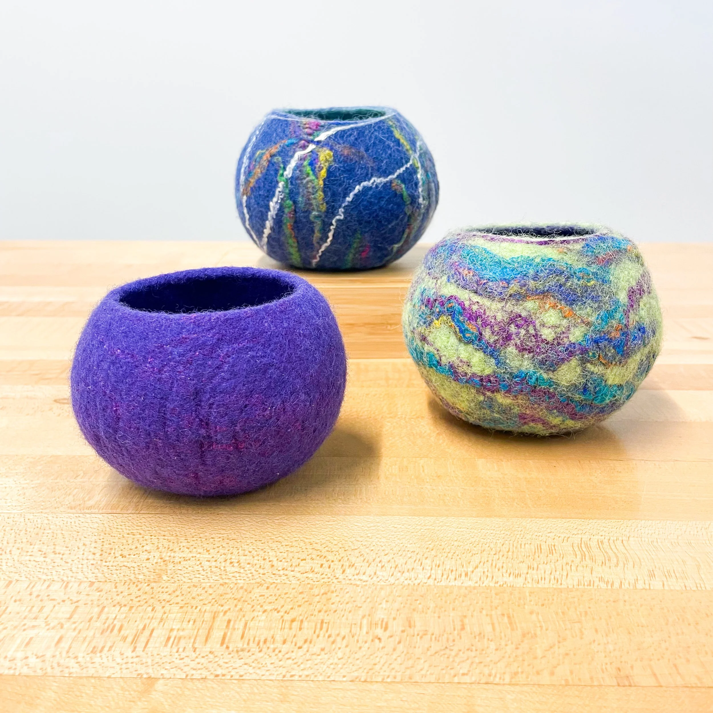 Three felt bowls, purple, shades of green, and blue with colorful accents sit on a light wood table.