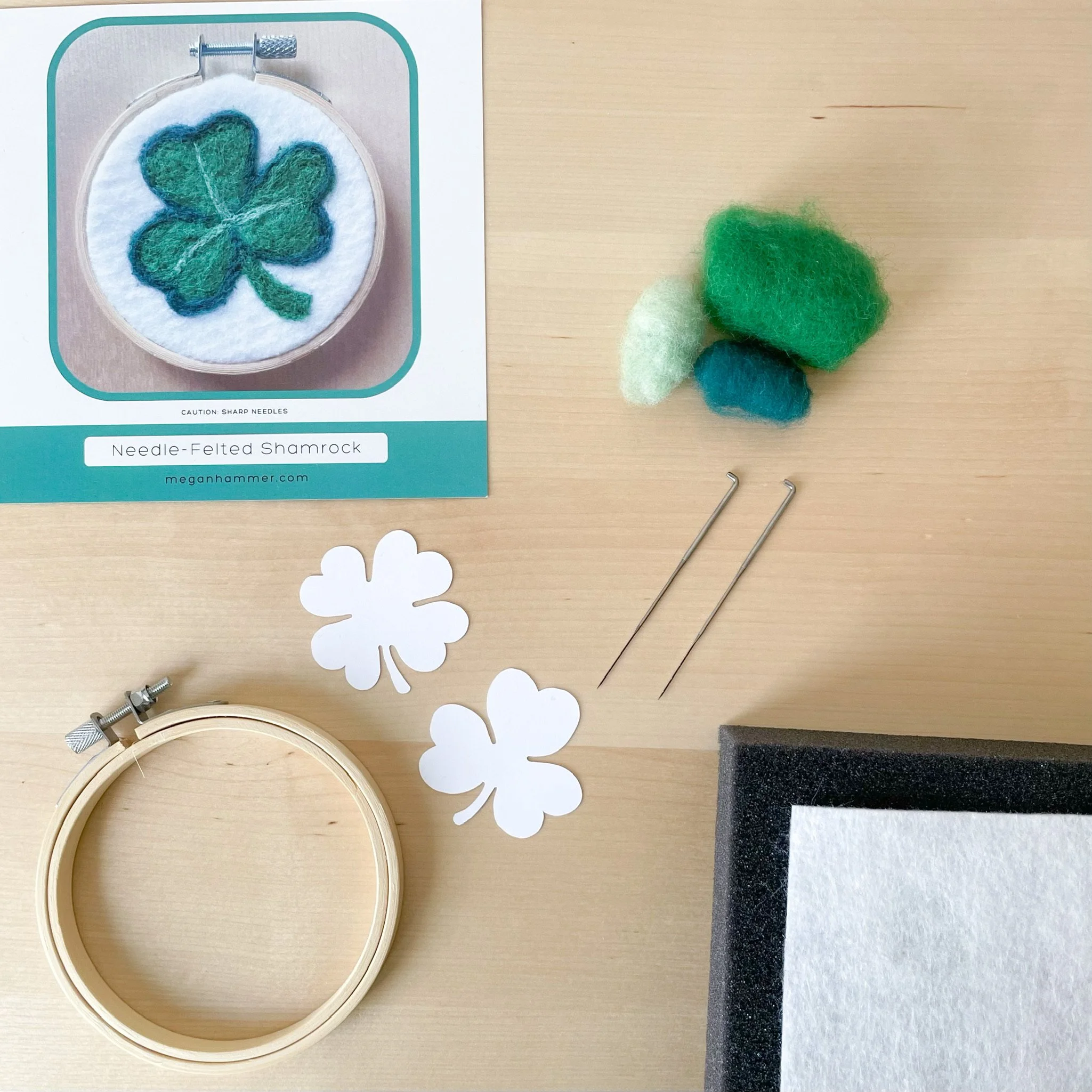 Kit tools and supplies laid out on a wood surface: shamrock label, tufts of colorful wool, stencils, hoop, two needles, black foam, and white felt.