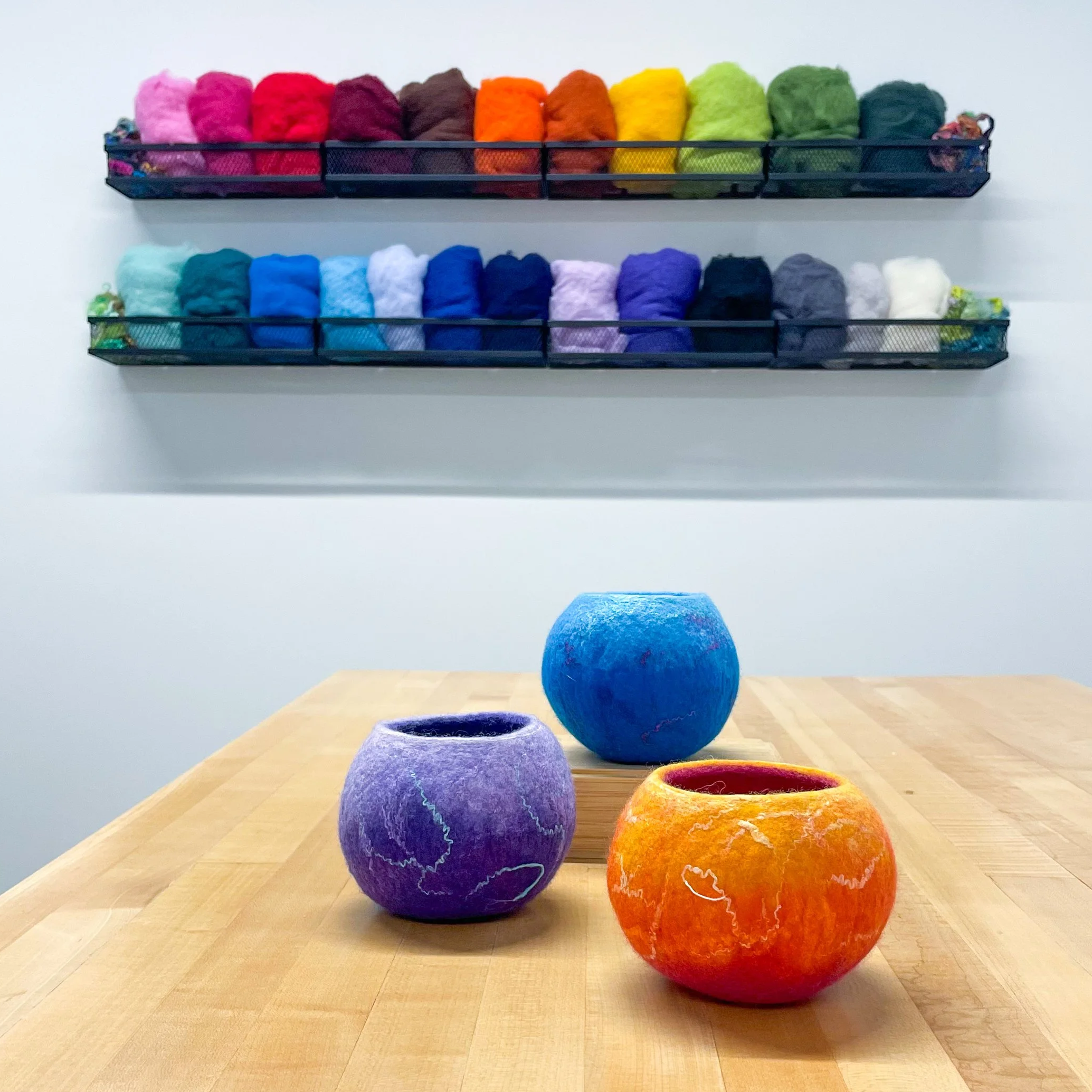 Three wet-felted bowls in blue, purple, and orange sit on a light wood table with a display of colorful wool on the wall behind.