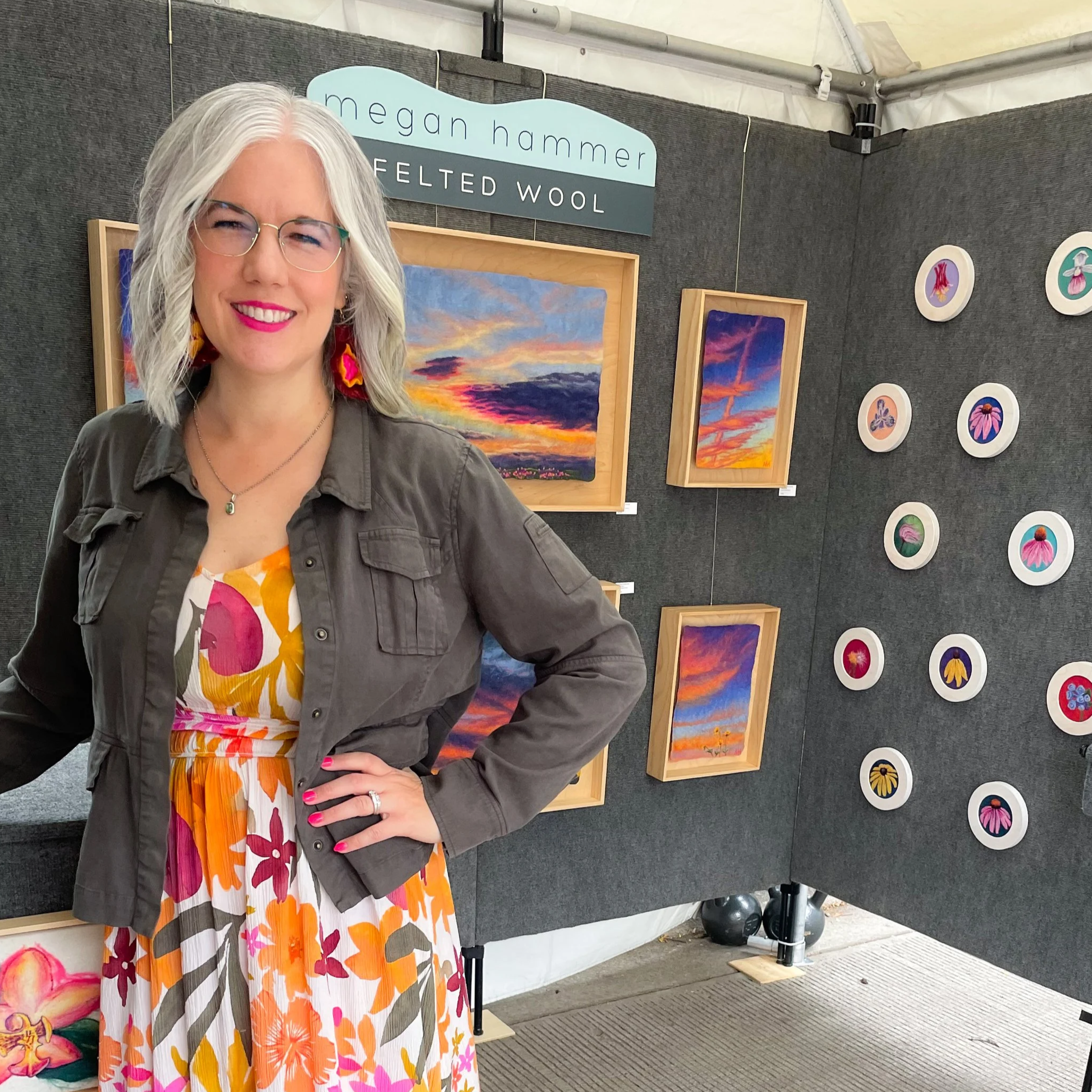 White woman in a green jacket and colorful floral dress stands in an art festival booth with gray walls and colorful felted artwork hanging behind her.