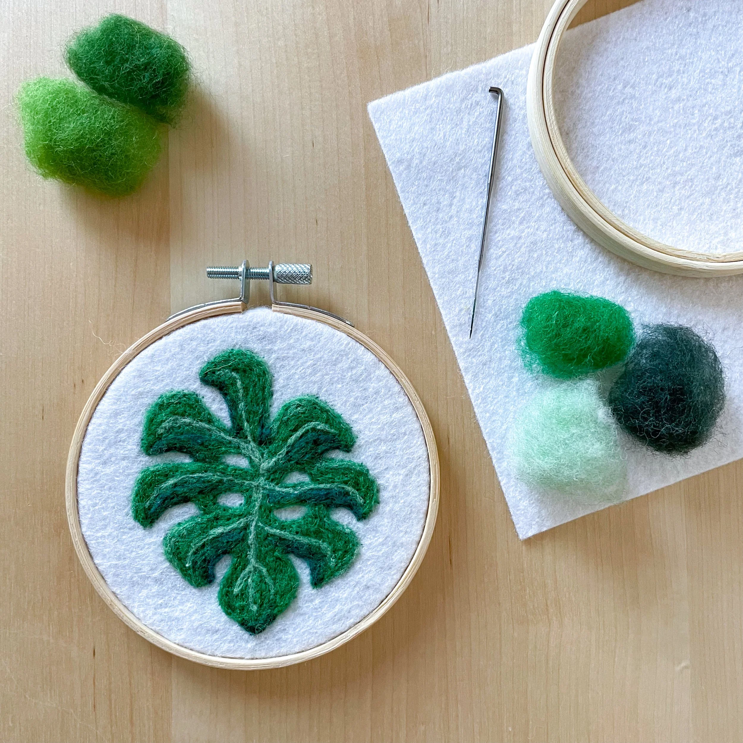 Fetled art of a monstera leaf framed in a hoop laying on a wood table. Tufts of colorful wool, white felt, a hoop, and a needle surround the art.