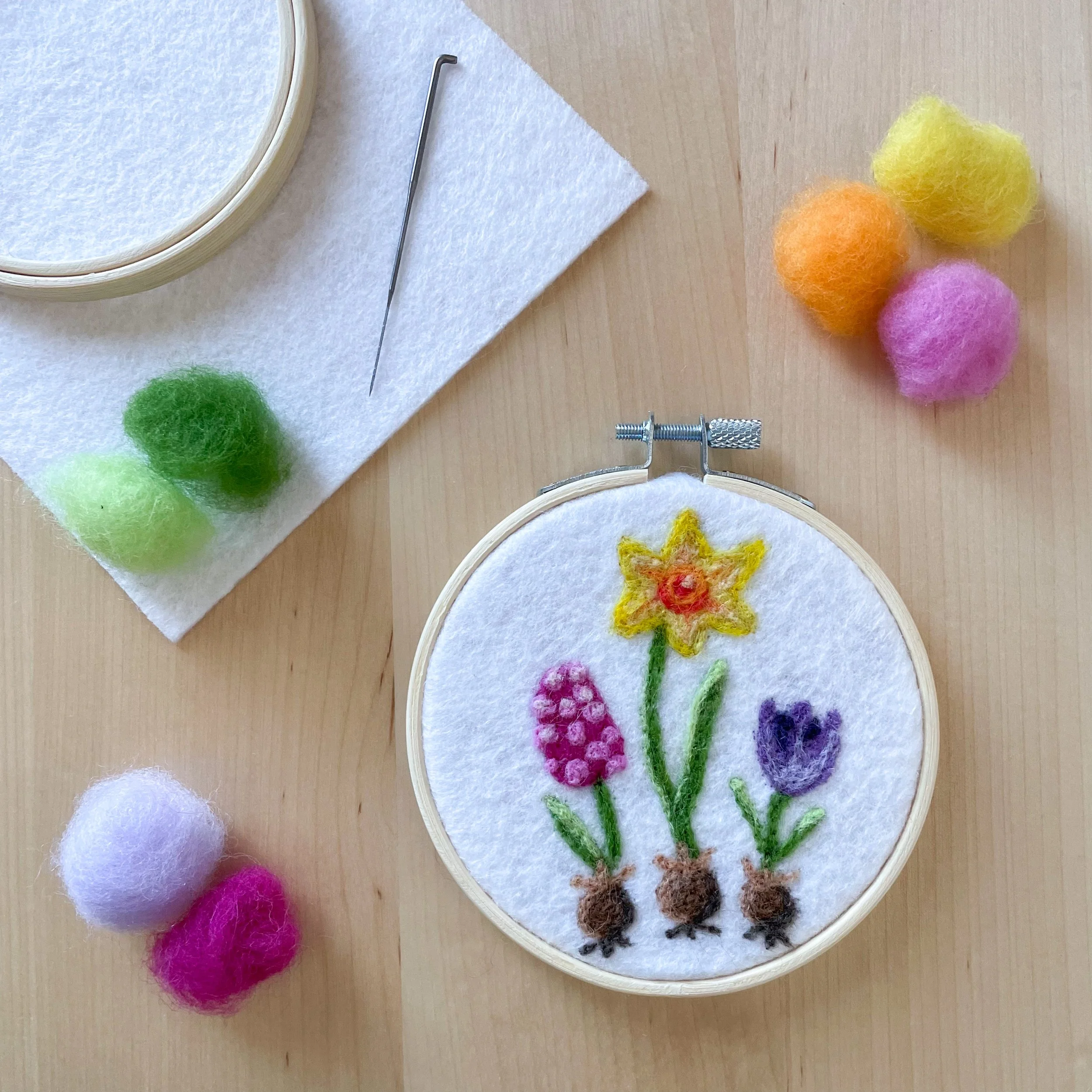 Felted art of three colorful flower bulbs framed in a hoop. Art is sitting on a wood table surrounded by colorful tufts of wool, white felt, a hoop, and a needle.