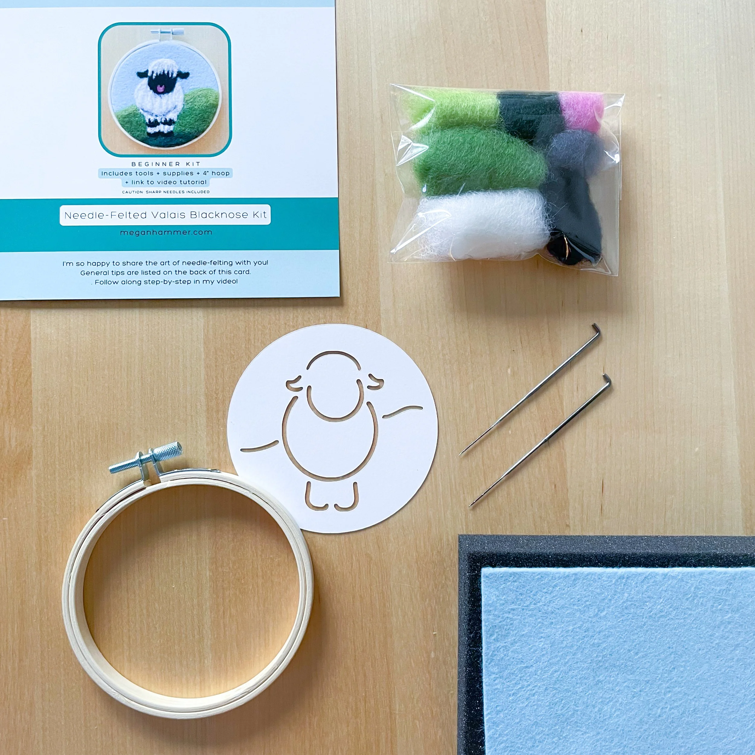 Kit tools and supplies laid out on a wood surface: Valais Blacknose Sheep label, packet of colorful wool, stencil, hoop, two needles, black foam, and white felt.