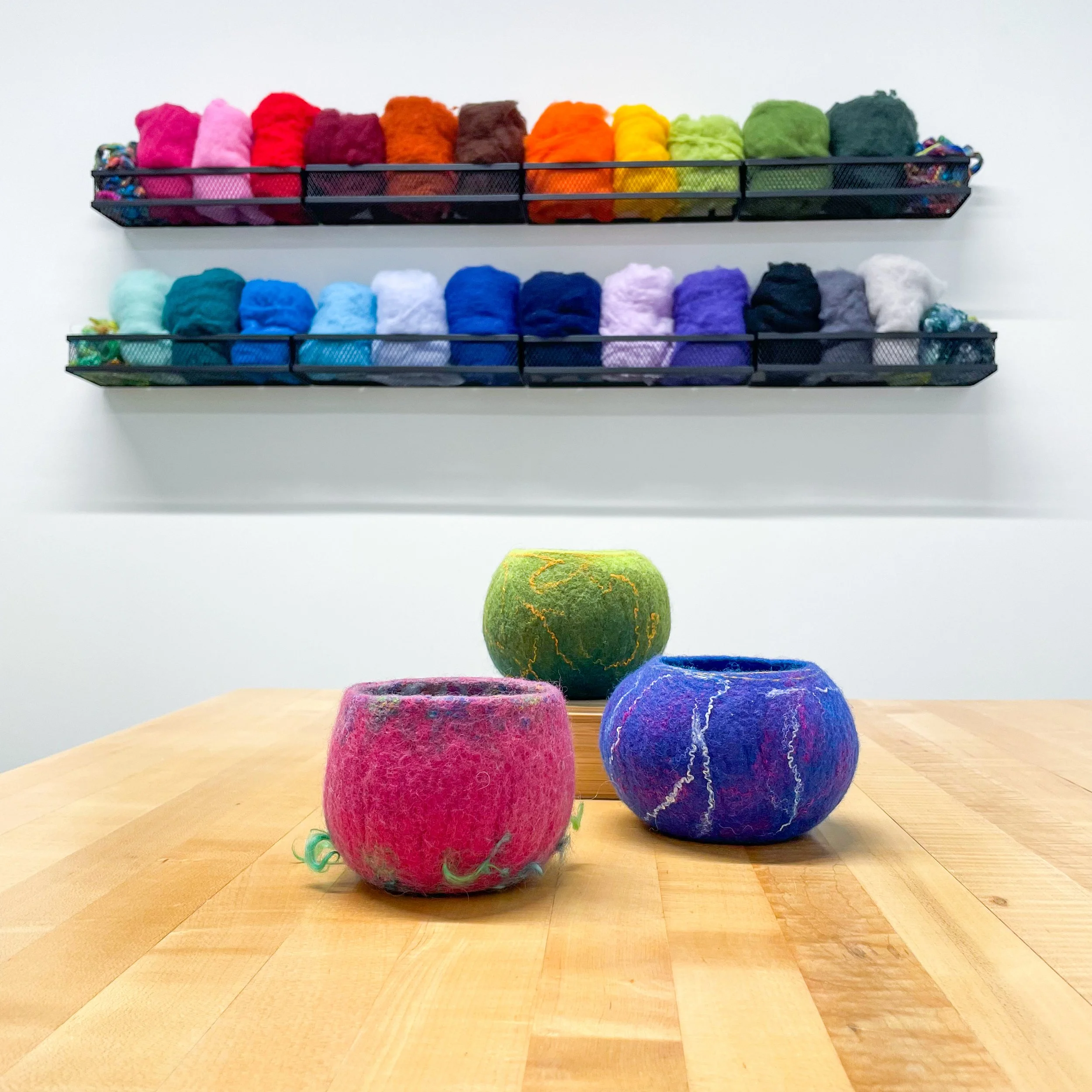 Three colorful felt bowls in shades of pink, green, and purple sit on a light wood table with a display of colorful wool on the wall behind.