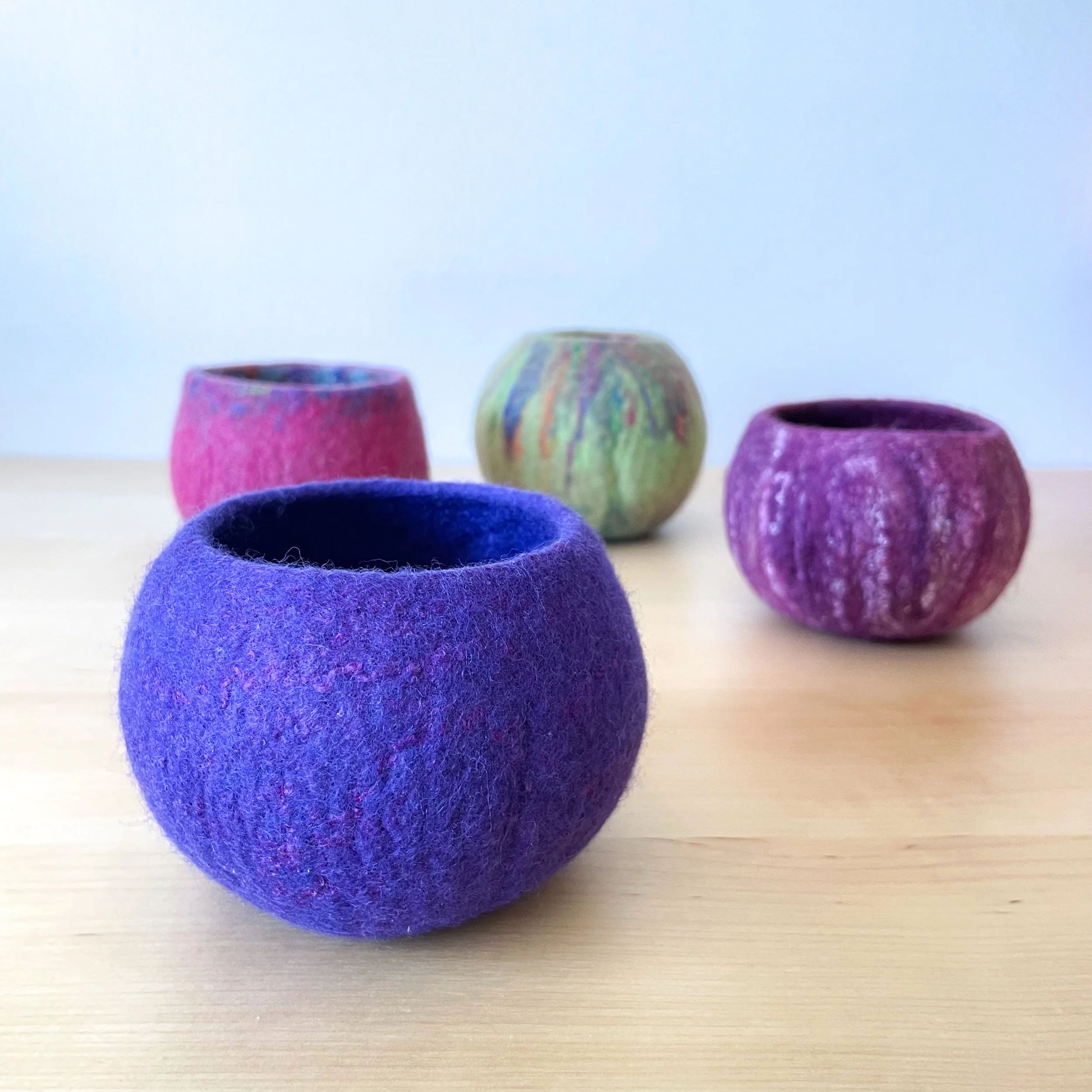 Four colorful felt bowls in shades of pink, green, and purple sit on a light wood table.