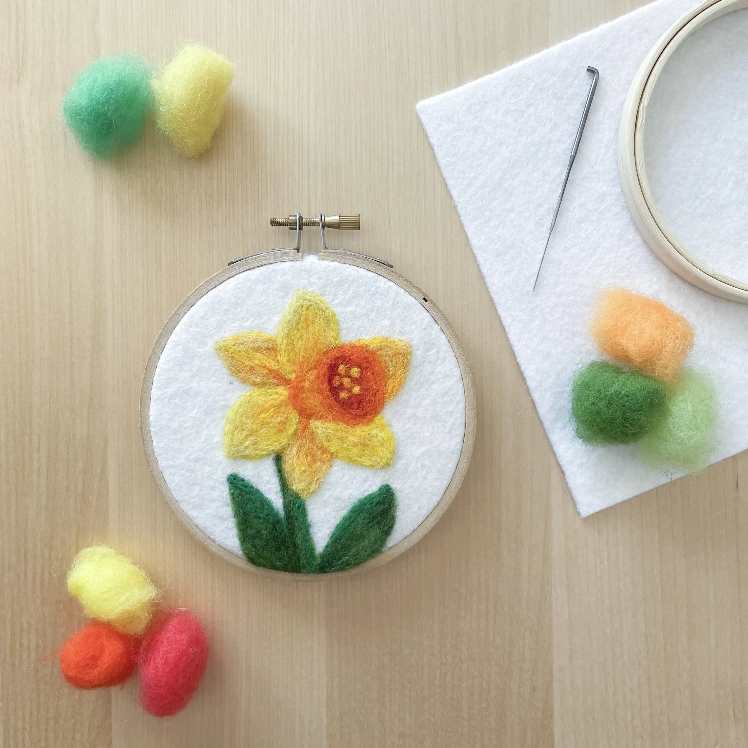 Felted art of a yellow flower and green stem and leaves framed in a hoop. Art is sitting on a wood table surrounded by colorful tufts of wool, white felt, a hoop, and a needle.