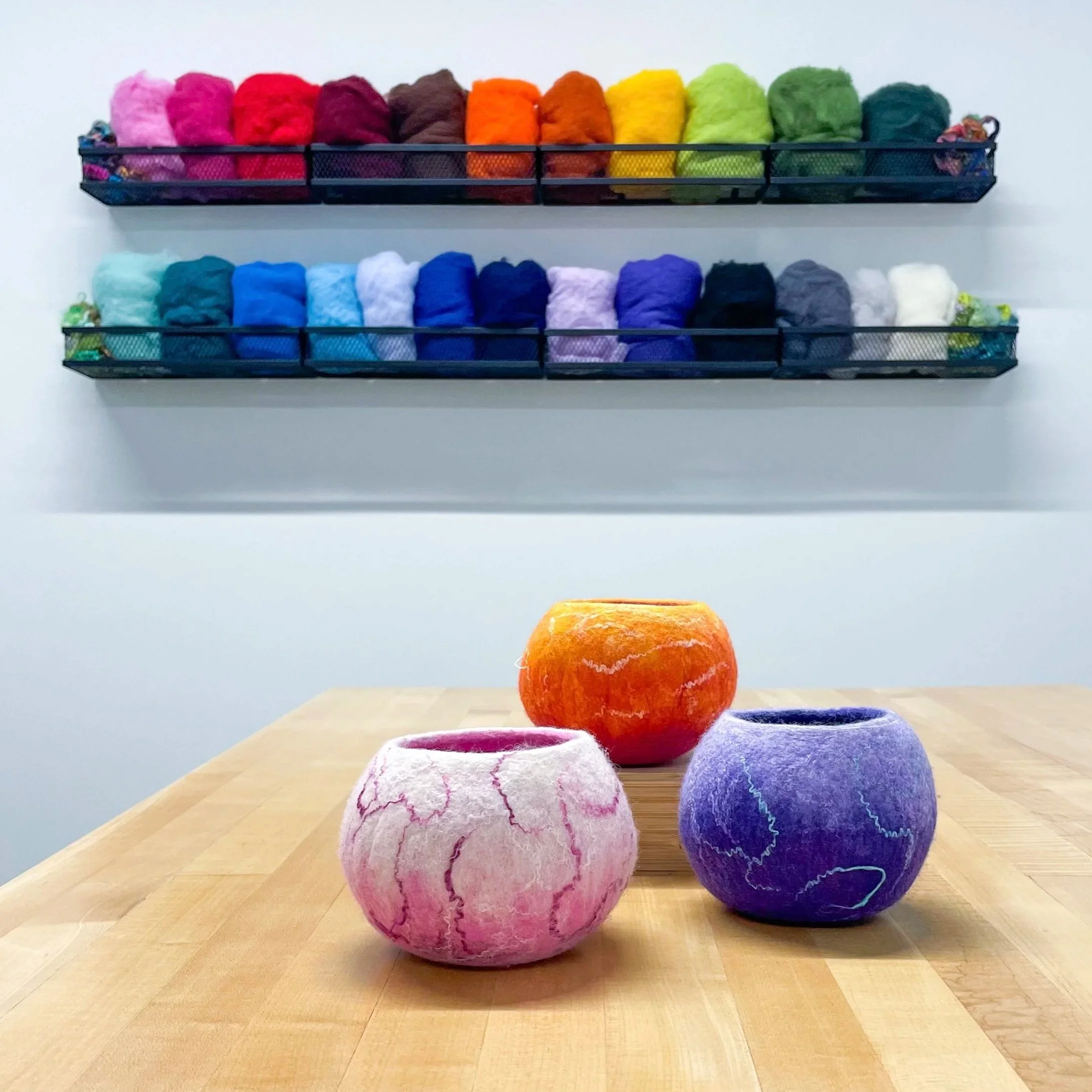 Three colorful felt bowls sit on a wood table with a colorful wool display on the wall behind. Wet-Felting workshop taught by Megan Hammer Art.