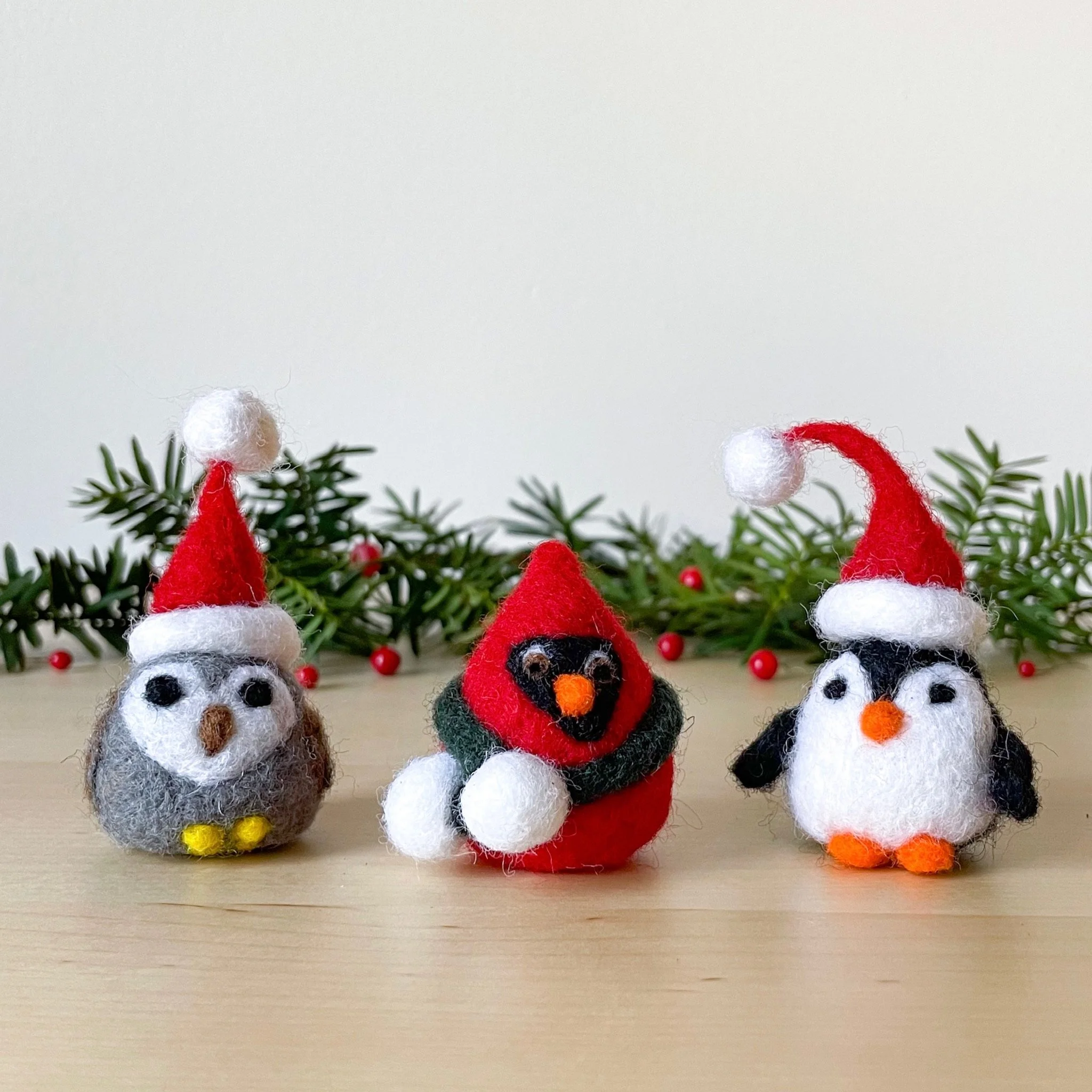 Three felted birds (gray owl and black and white penguin in red Santa hats, and a red cardinal with a green scarf)  sit on a wood table with pine boughs and red berries behind them.