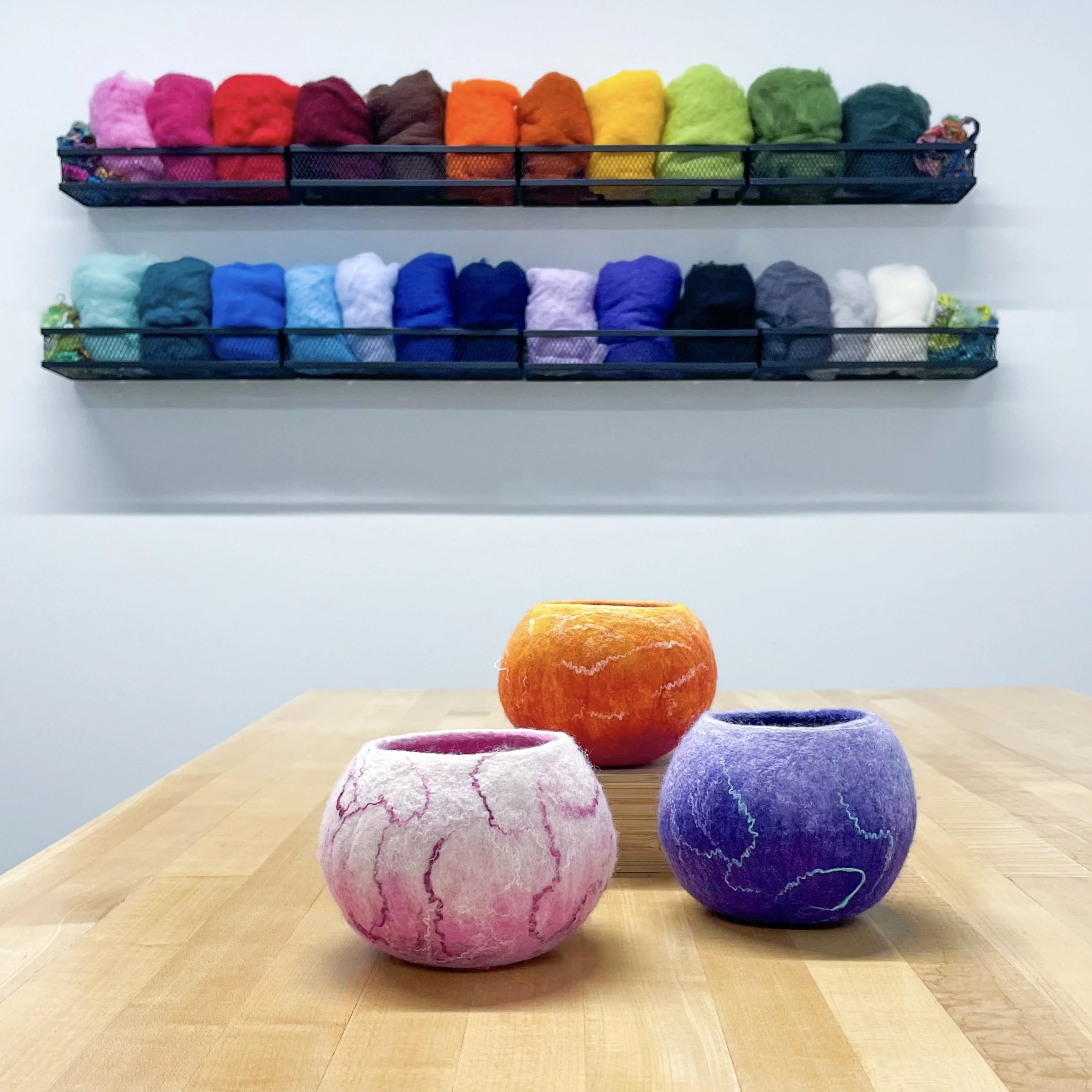 Three colorful felt bowls in shades of pink, orange, and purple sit on a light wood table with colorful wool displayed on the wall behind.